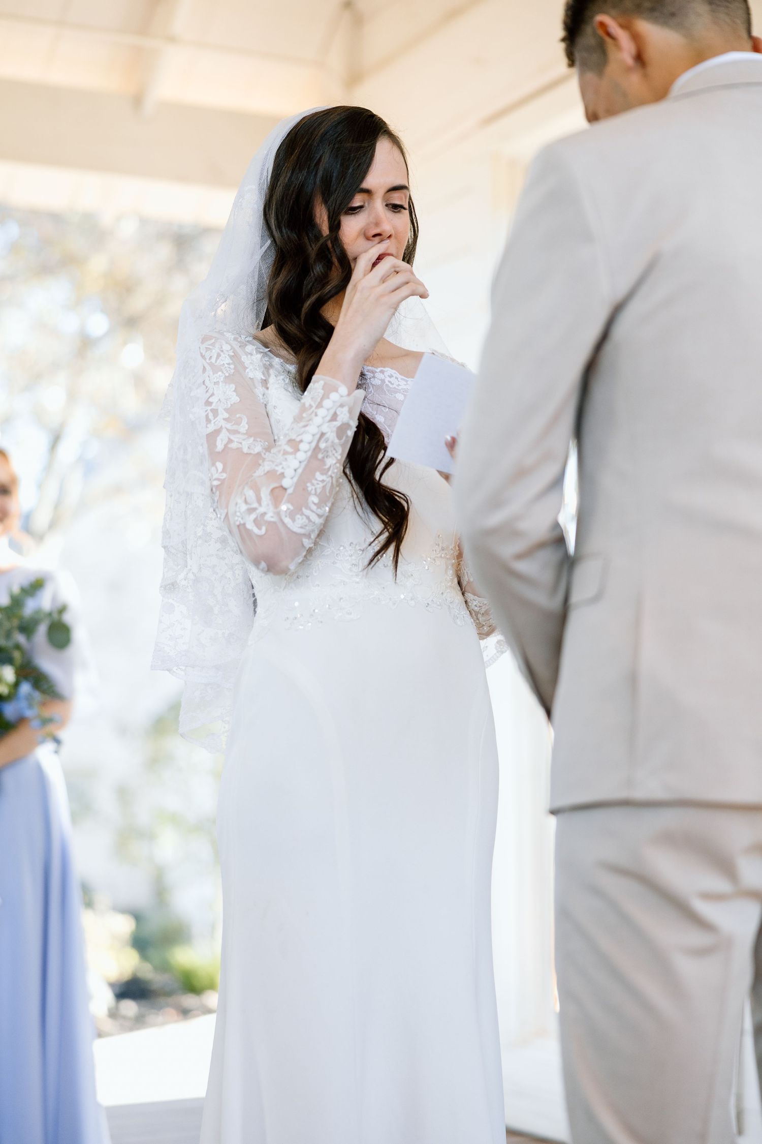 Tearful bride in lace veil reading wedding vows during outdoor ceremony.