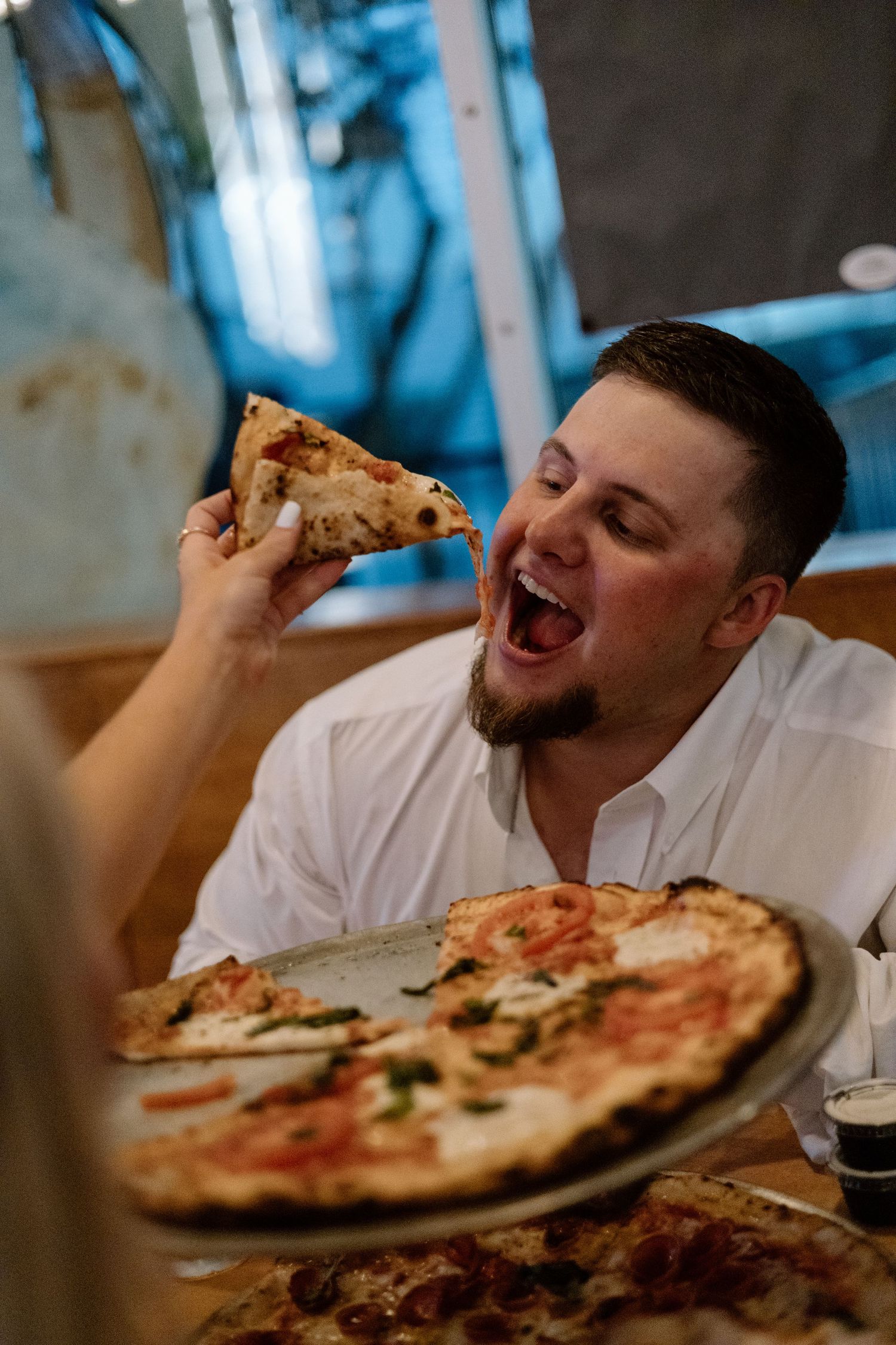 Man in white shirt excitedly eating a slice of Neapolitan pizza at a restaurant table.