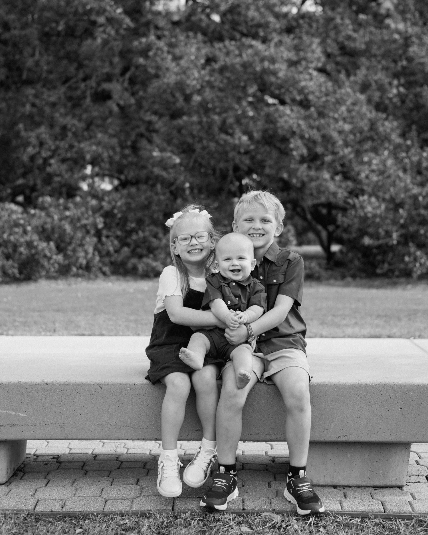 Three young children sitting together on a bench outdoors, smiling, in a black and white photo.