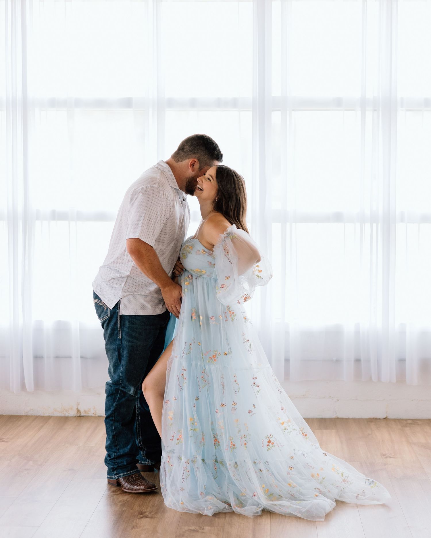 Couple sharing an intimate moment in a bright studio, woman in a floral light blue gown, man in white shirt and jeans.