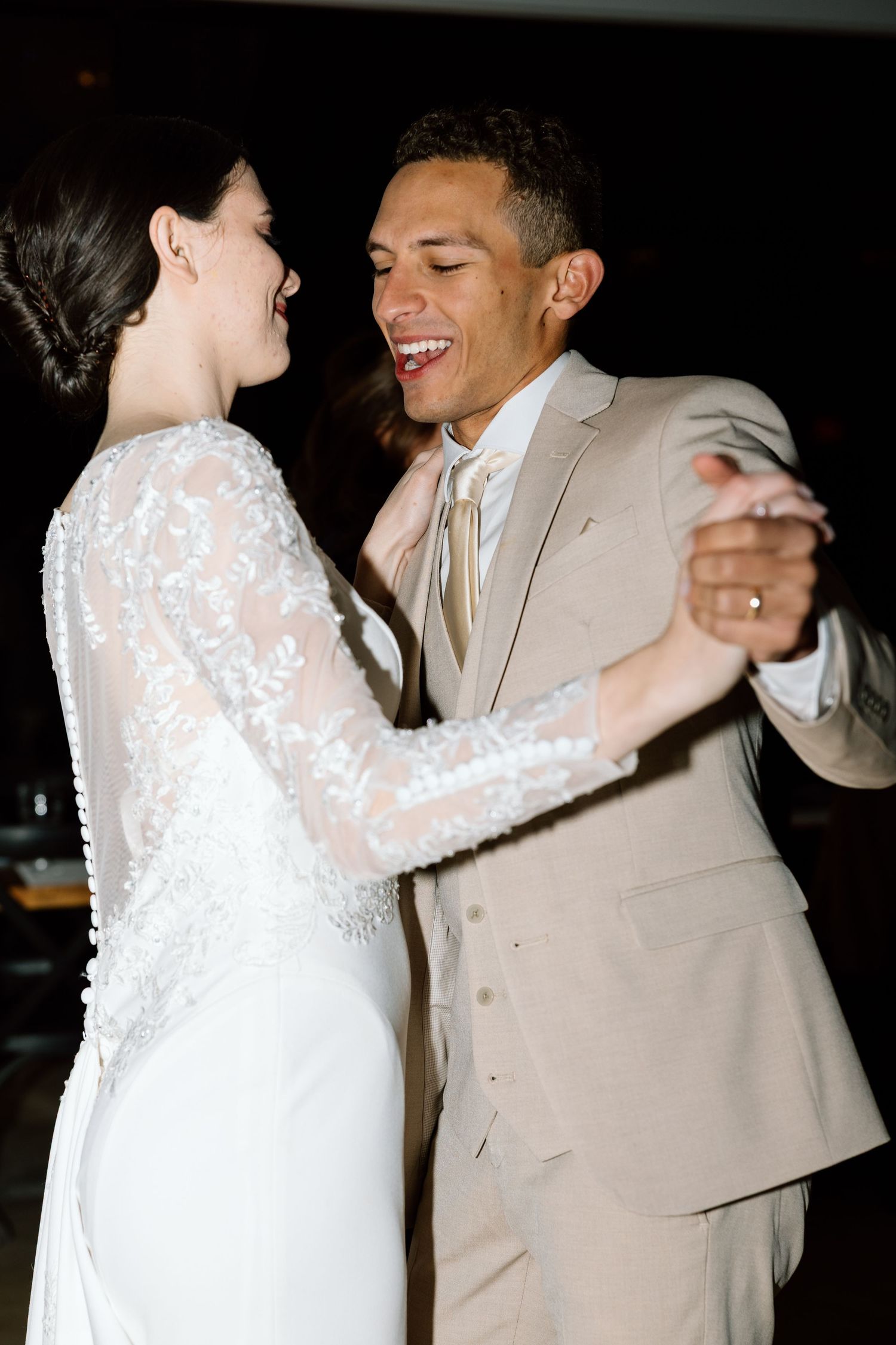Bride and groom share their first dance, smiling joyfully, she in lace gown, he in tan suit with ivory tie.