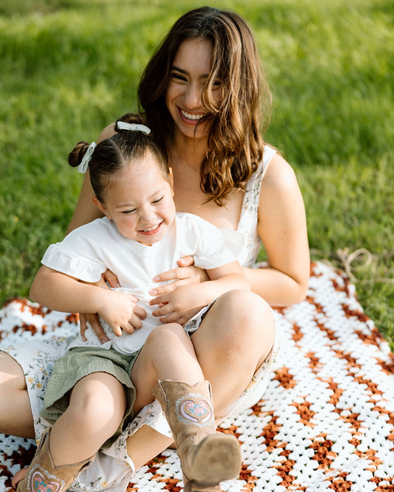 A smiling mother and young daughter laugh together on a crocheted blanket in a sunny green park.