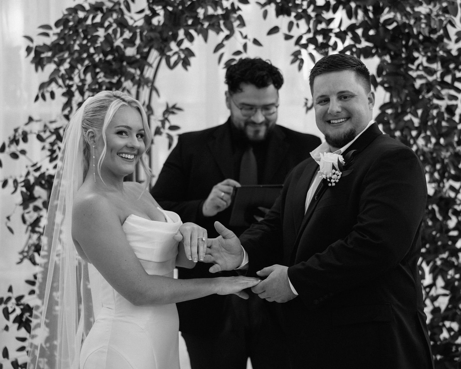 Black and white photo of smiling bride and groom exchanging rings during wedding ceremony with officiant.