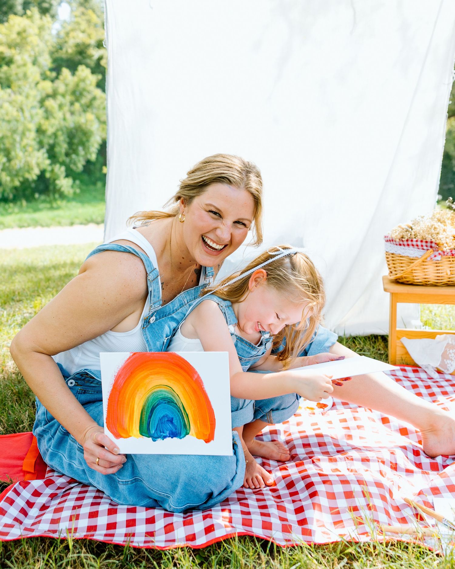 Woman and young girl sitting on red checkered blanket outdoors, smiling and holding a colorful rainbow painting.