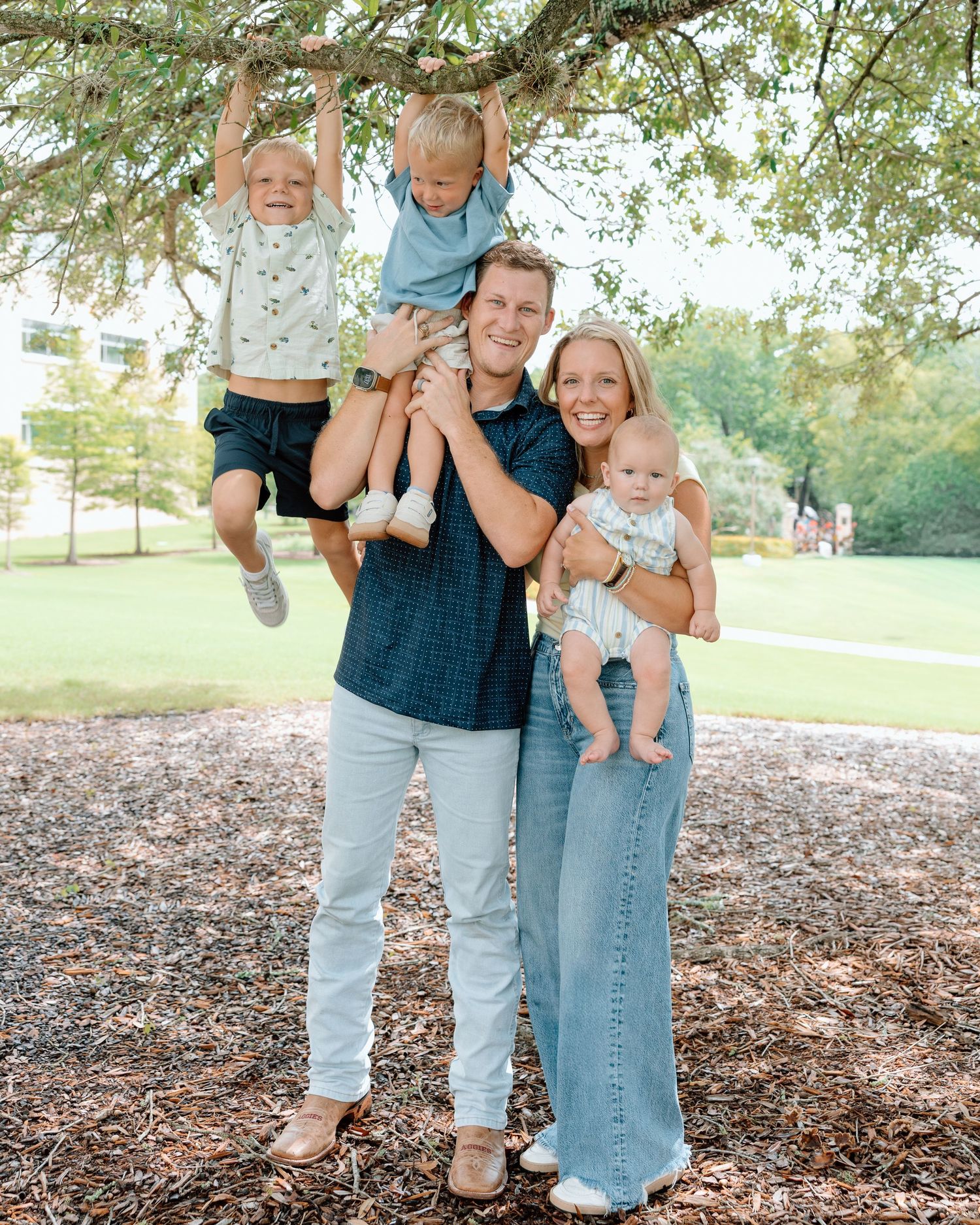 Happy family of five posing outdoors under a tree, parents holding three young children in a sunny park setting.