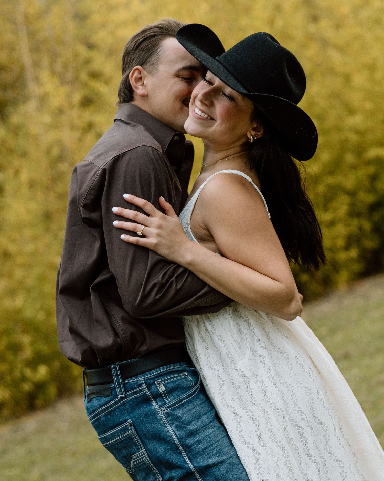 Couple embracing and smiling in autumn setting, woman wearing black cowboy hat and engagement ring.