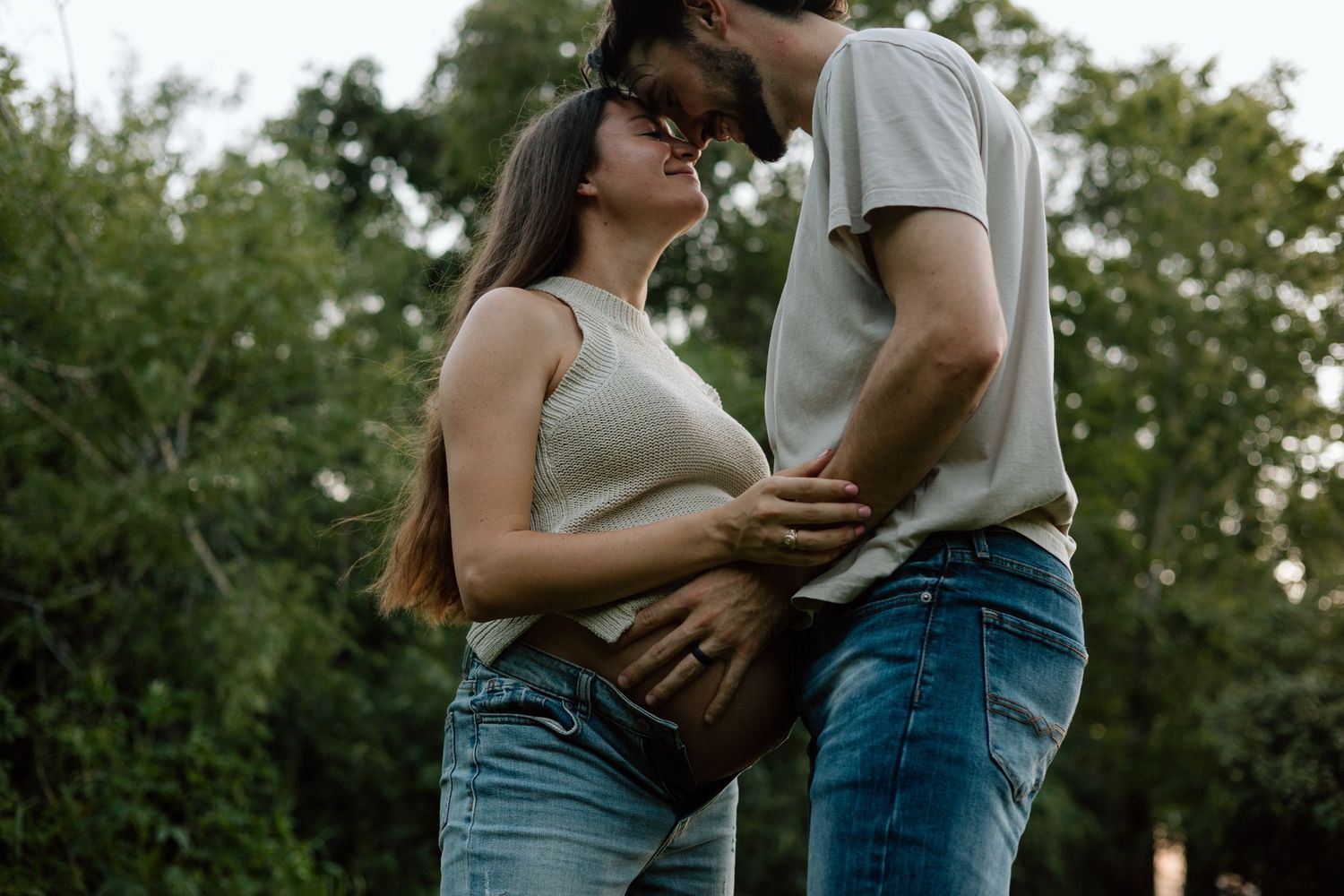 A tender moment between an expecting couple in casual attire standing in a green outdoor setting.