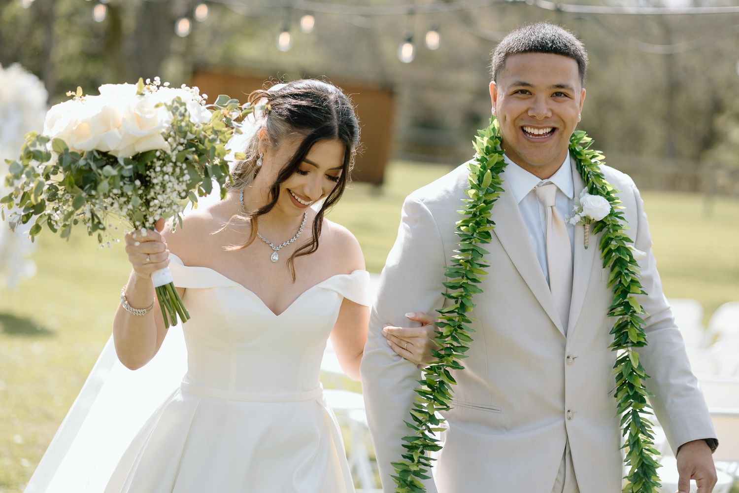 Happy newlyweds walk together outdoors wearing white wedding attire and green leaf leis.