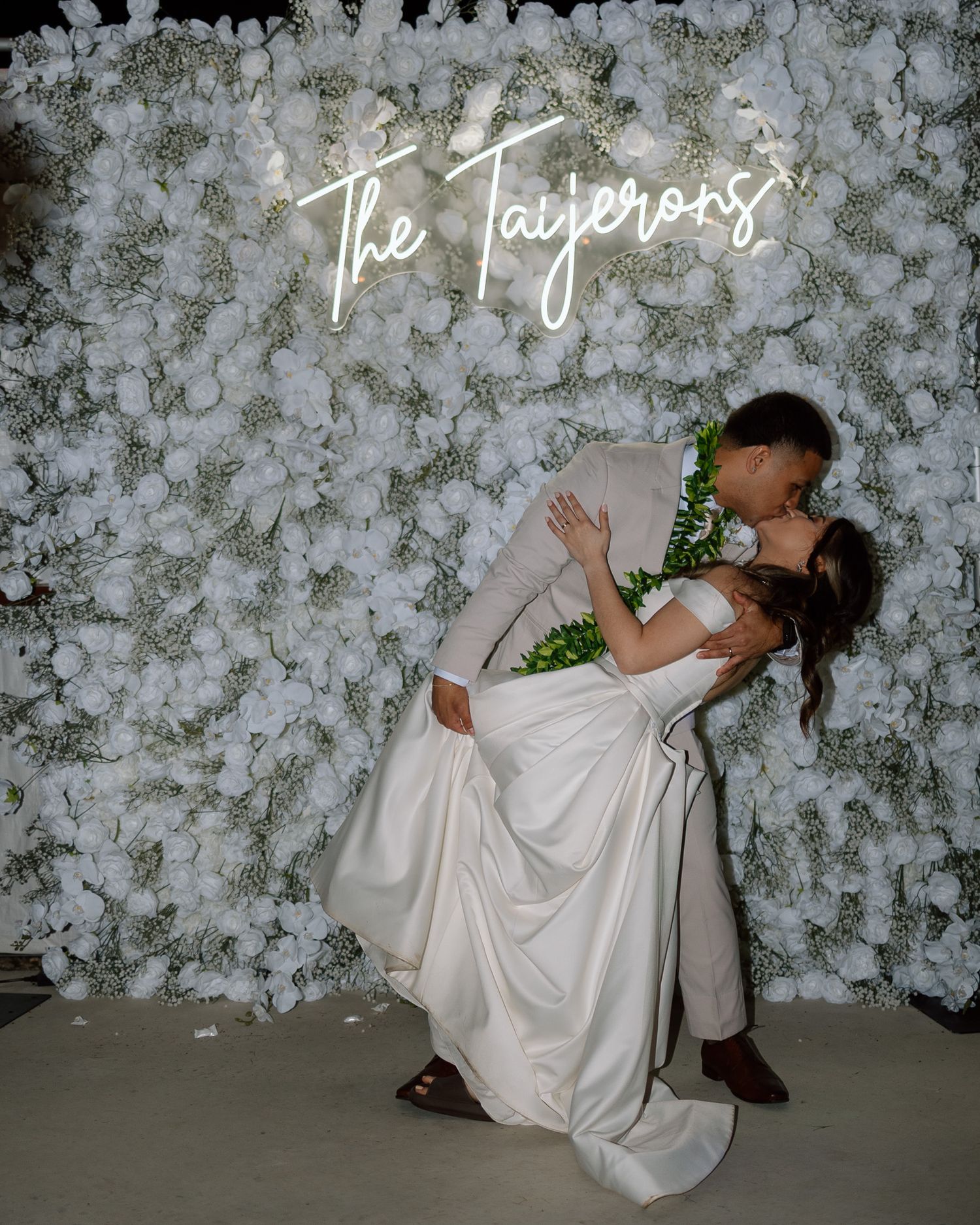 Romantic dip pose in front of illuminated floral wall at wedding reception.