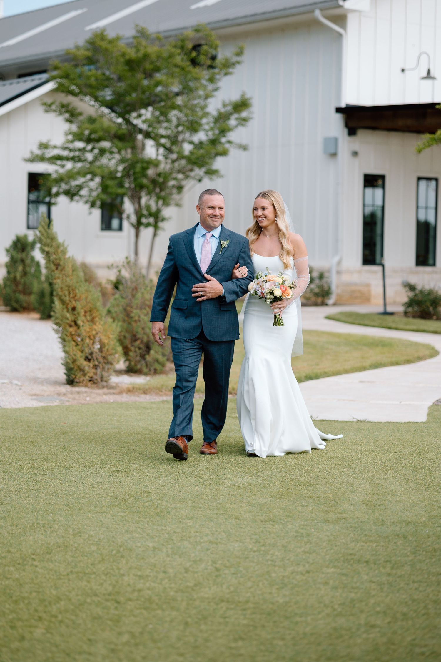 A walk along a white modern venue's grassy path with wedding bouquet.