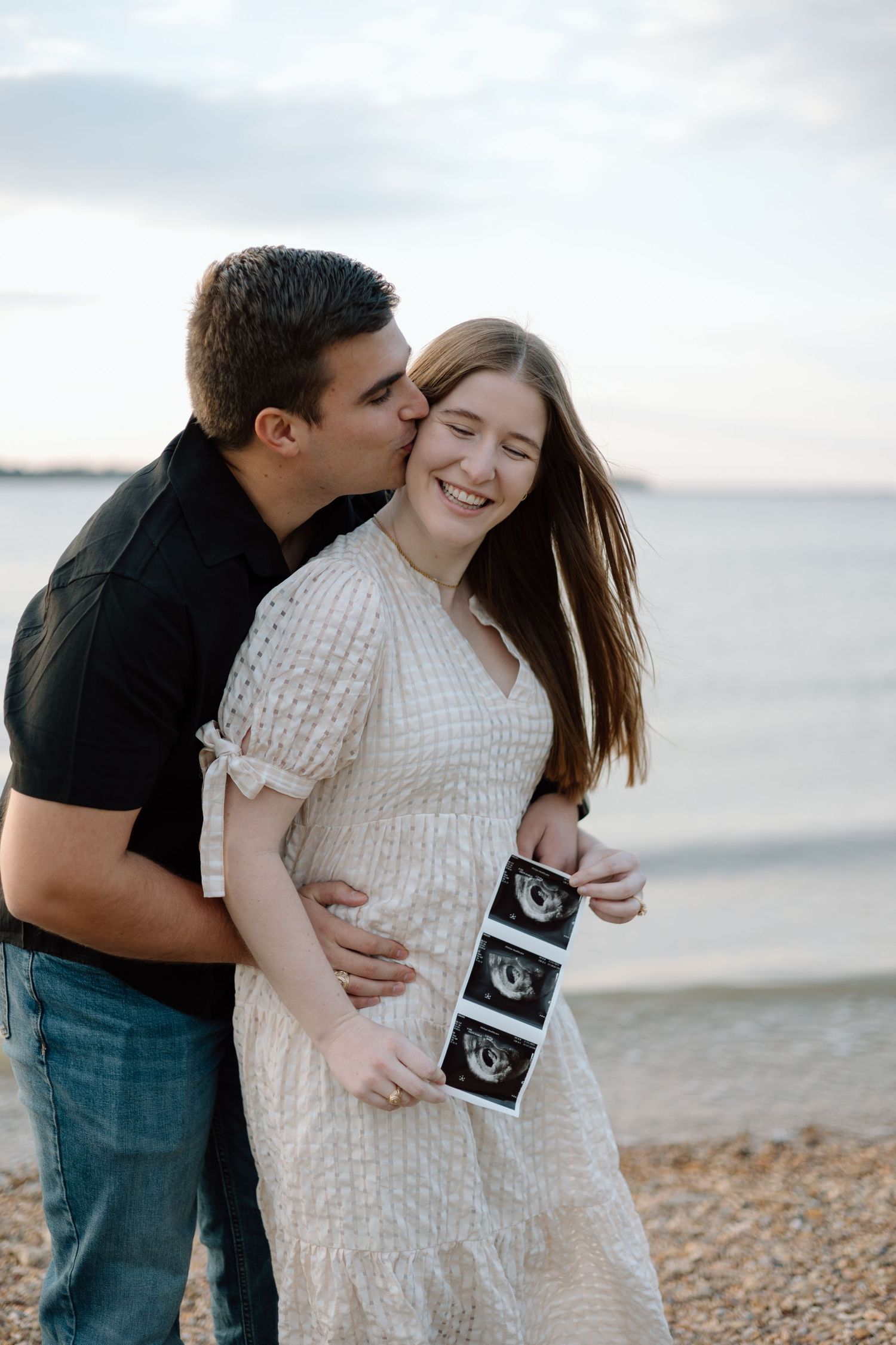 A couple shares a romantic moment on a beach while holding ultrasound photos.