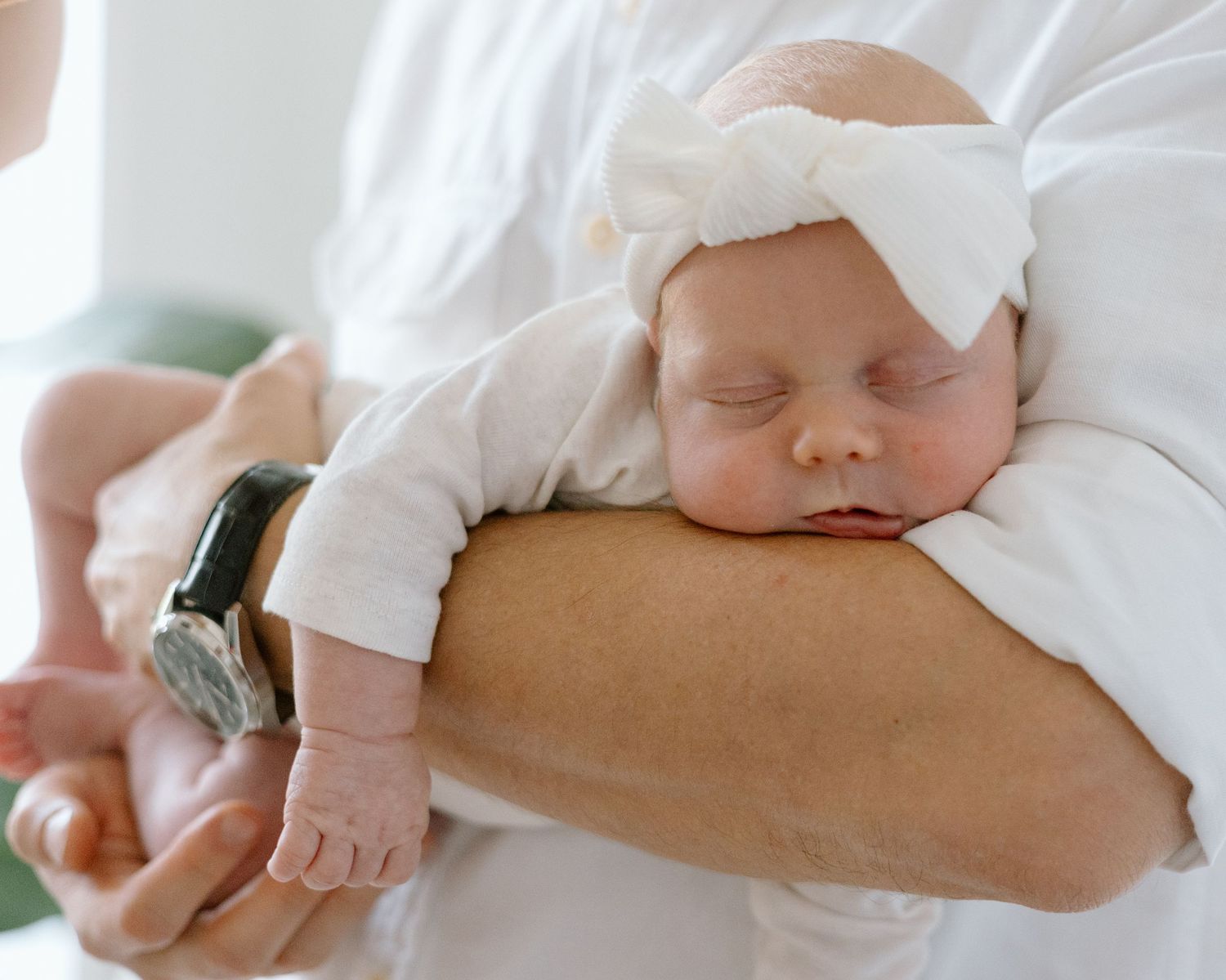 Sleeping newborn in white outfit with bow headband rests peacefully on caregiver's arm.