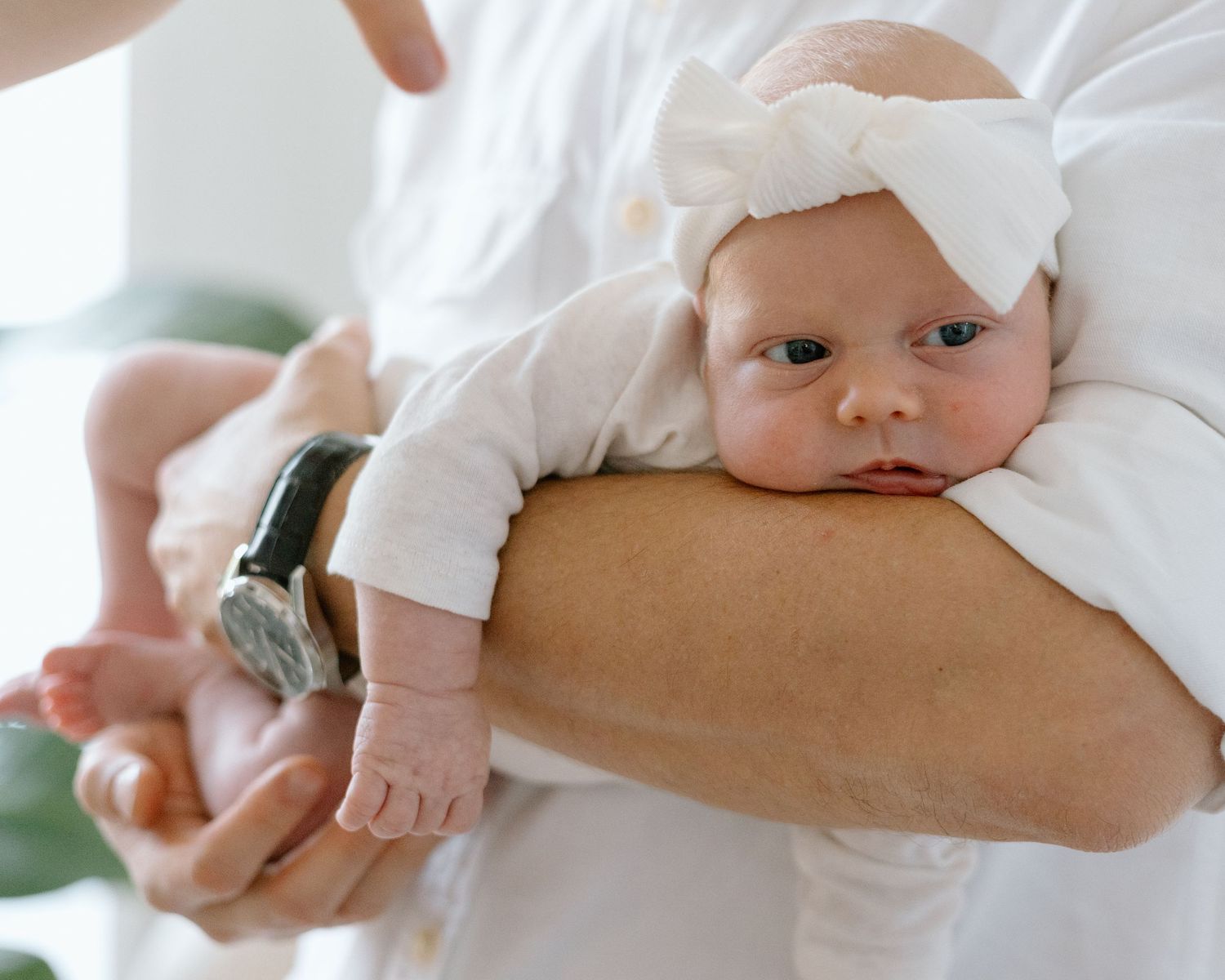 Alert baby in white outfit with bow headband rests on caregiver's arm.