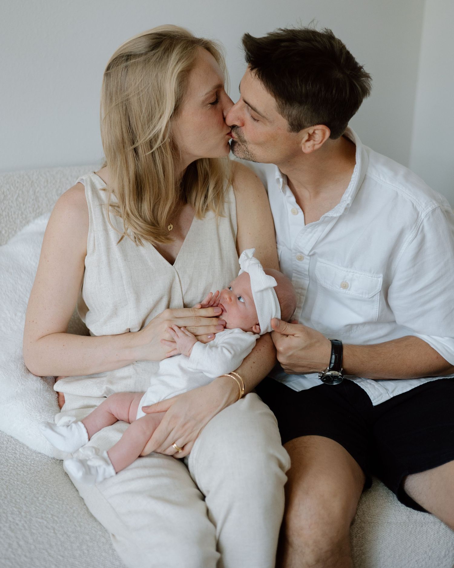 Intimate moment between couple holding their newborn baby on white couch in casual summer attire.