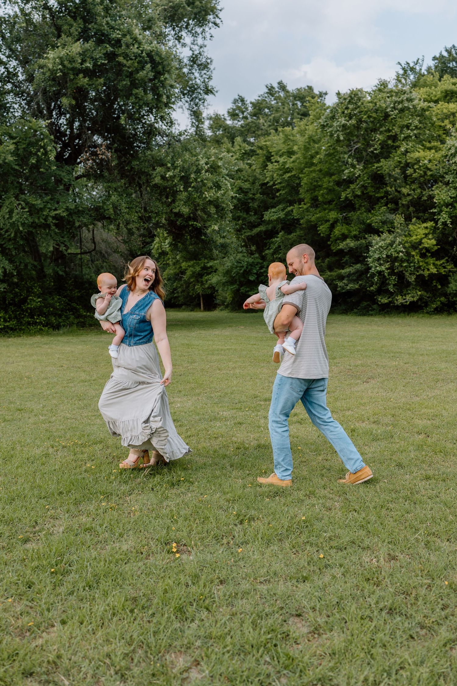 Adults and babies enjoying a carefree moment dancing together on green grass in a park setting surrounded by trees.