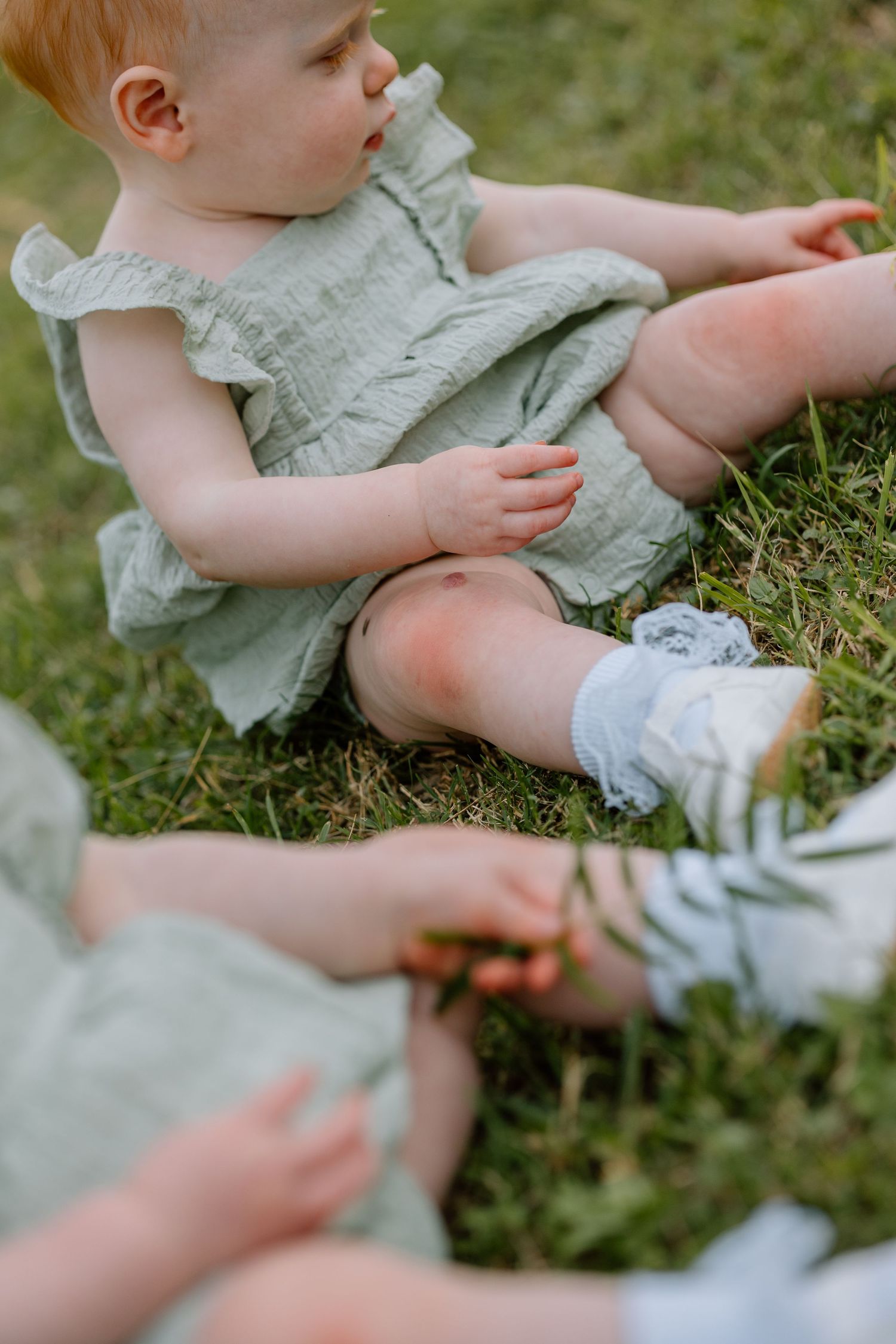 A young child in a ruffled mint dress sits on grass while reaching for something out of frame.