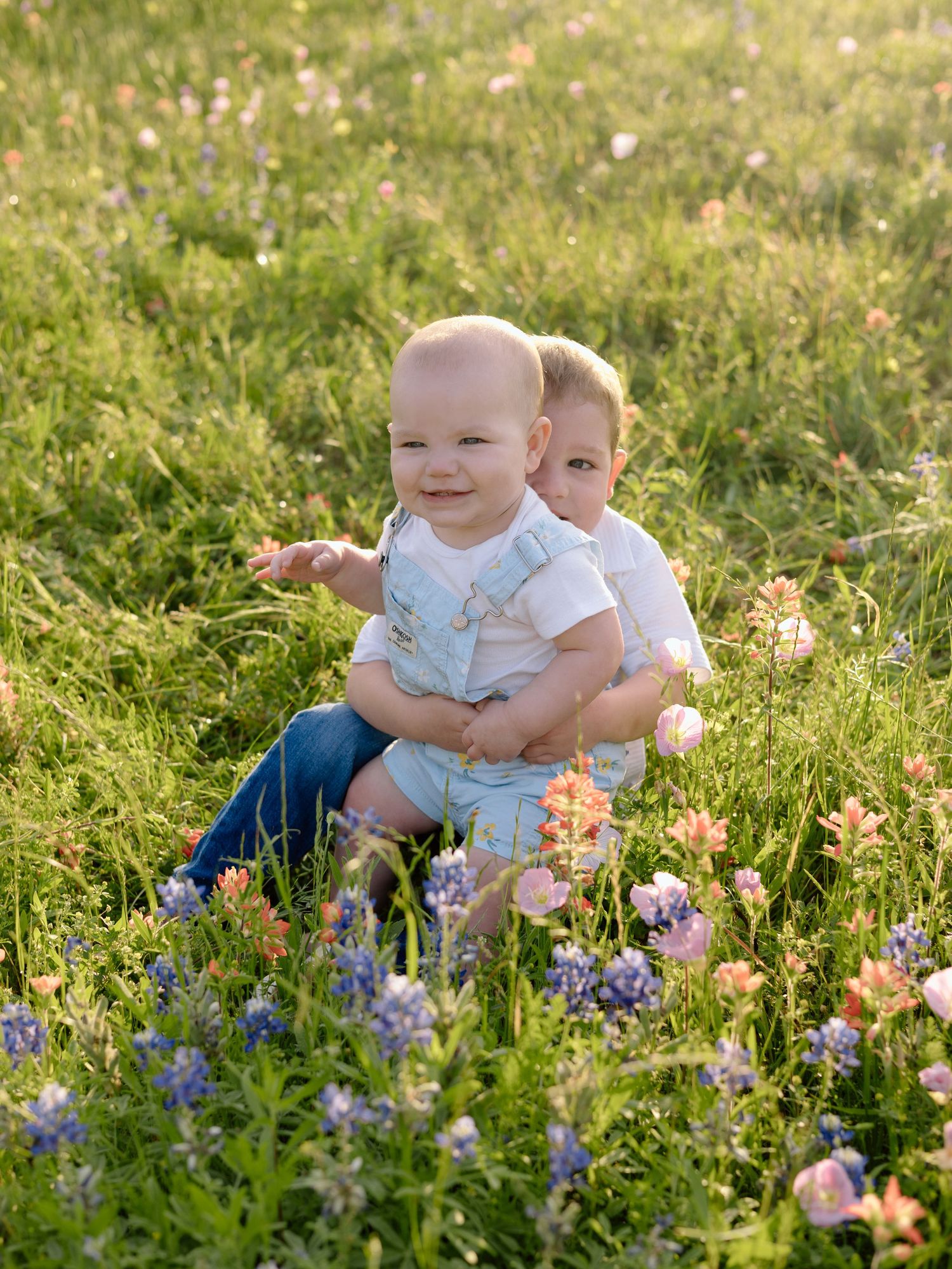 Two young children sit together in a sunny meadow filled with colorful wildflowers and soft golden light.