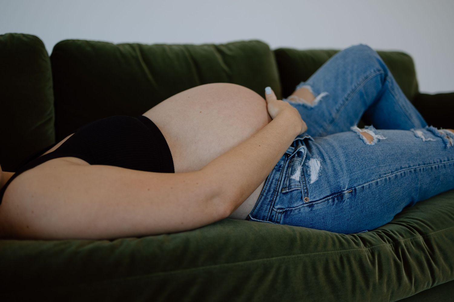 Someone wearing blue jeans and a black top lying on a dark green velvet couch.