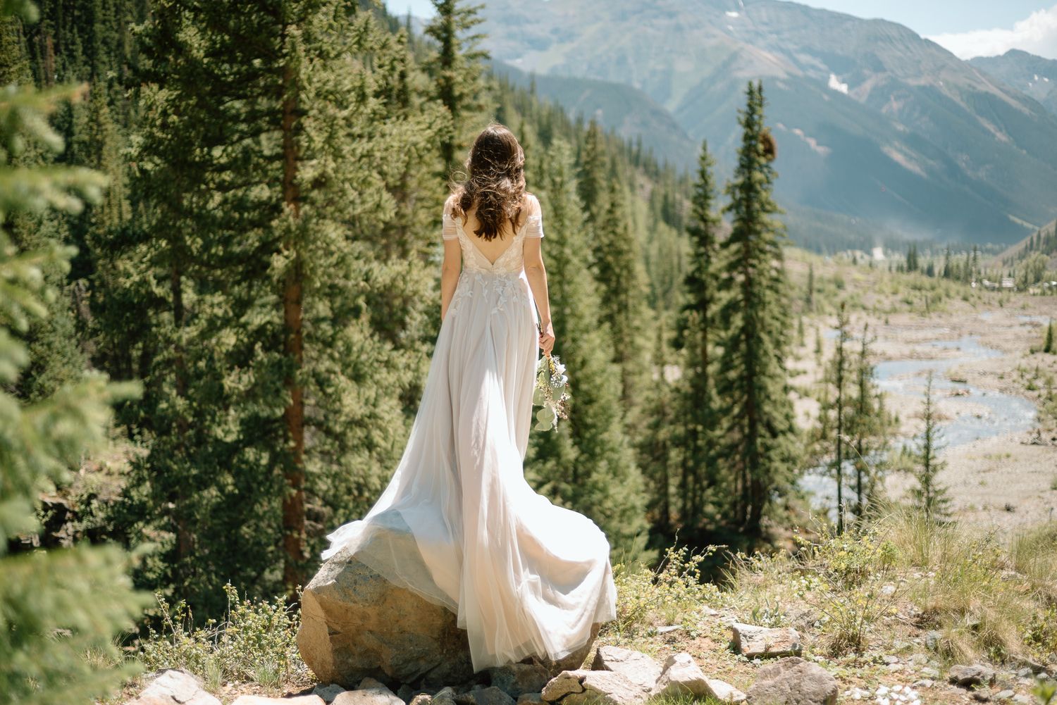 A bride in a flowing white dress stands on rocks against a mountain backdrop.