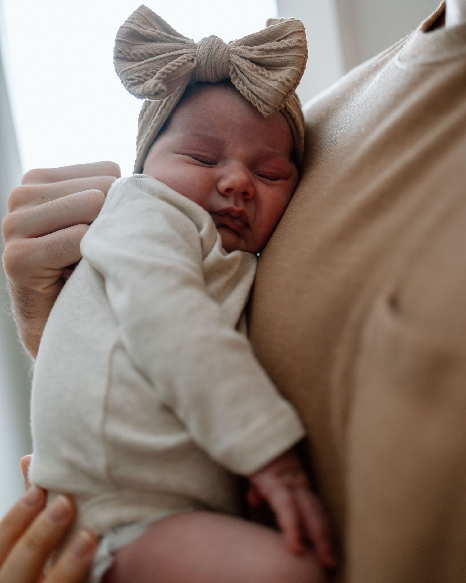 Sleeping newborn wearing beige outfit and bow headband rests against caregiver.