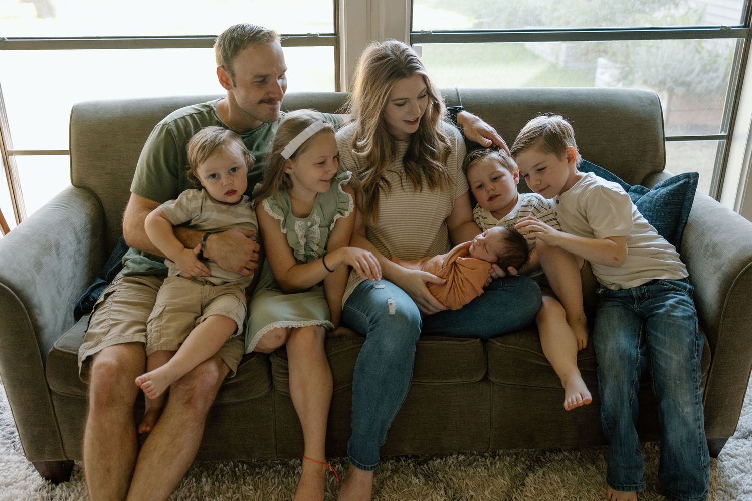 A happy family of six snuggled together on a gray couch in their living room while laughing and embracing each other.