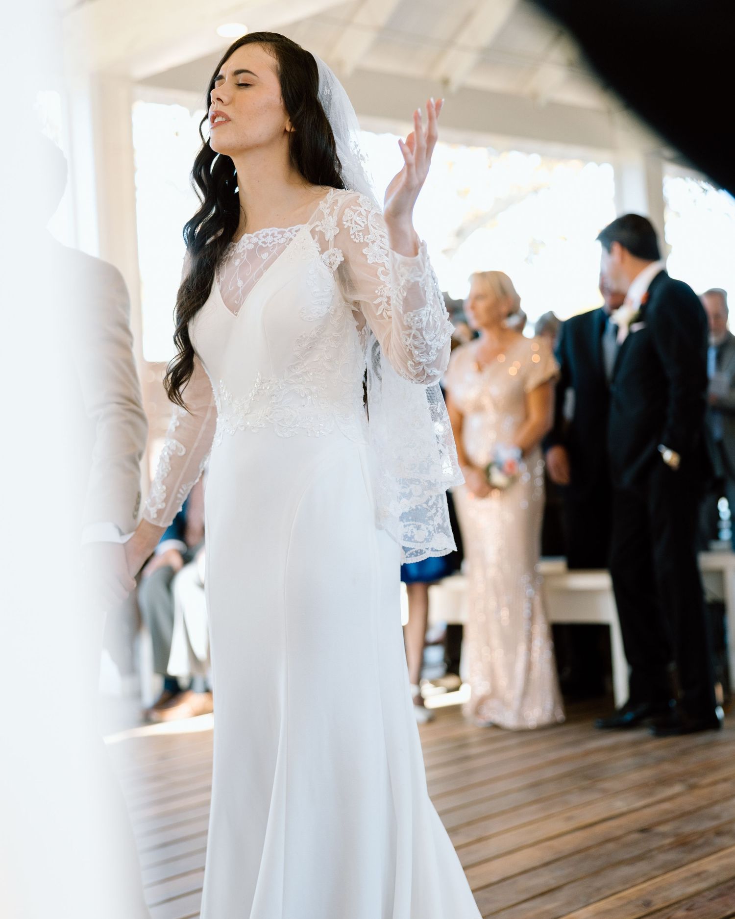 Dark-haired bride in lace sleeve wedding gown with veil during outdoor ceremony.
