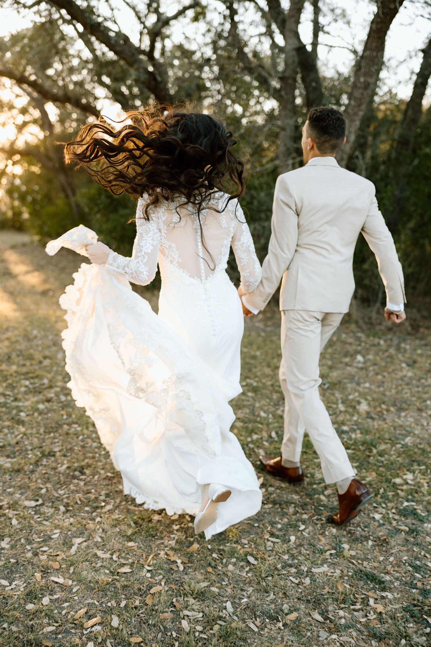 Bride and groom holding hands running through a sunlit outdoor path, bride in lace gown with flowing hair.