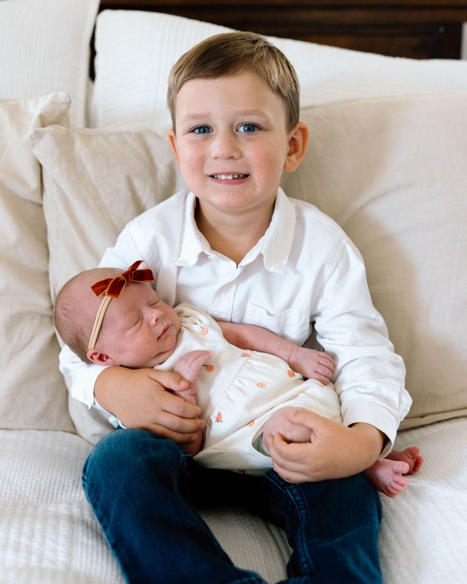 Young boy with blue eyes in white shirt holds sleeping newborn baby girl with red bow on a cream couch.