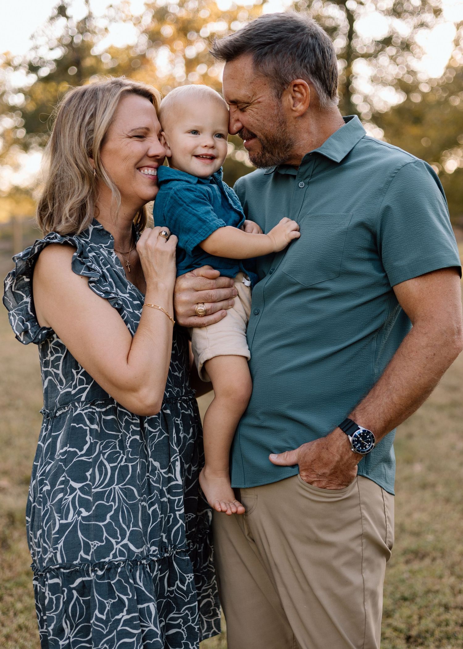 A smiling family with parents holding their happy baby boy outdoors in a sunlit field during a portrait session.