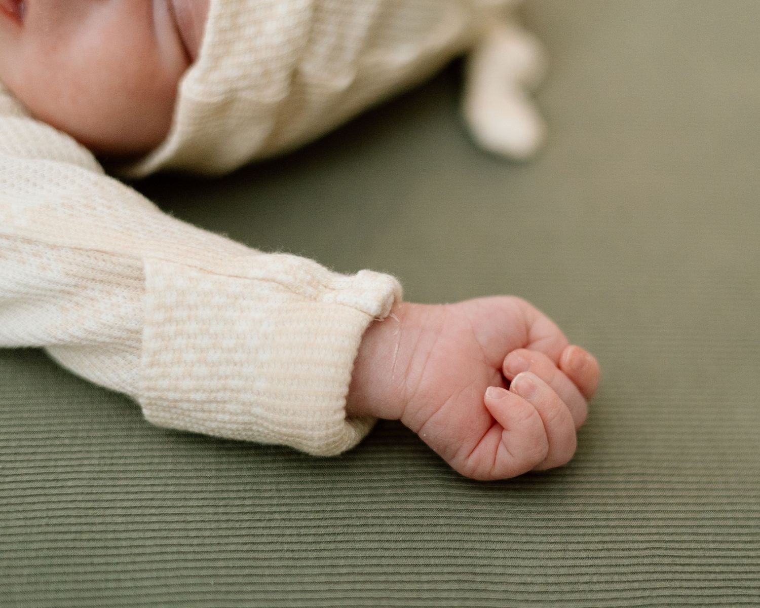 Close-up of a newborn baby's tiny clenched fist in a cream knit outfit, resting on a green surface.