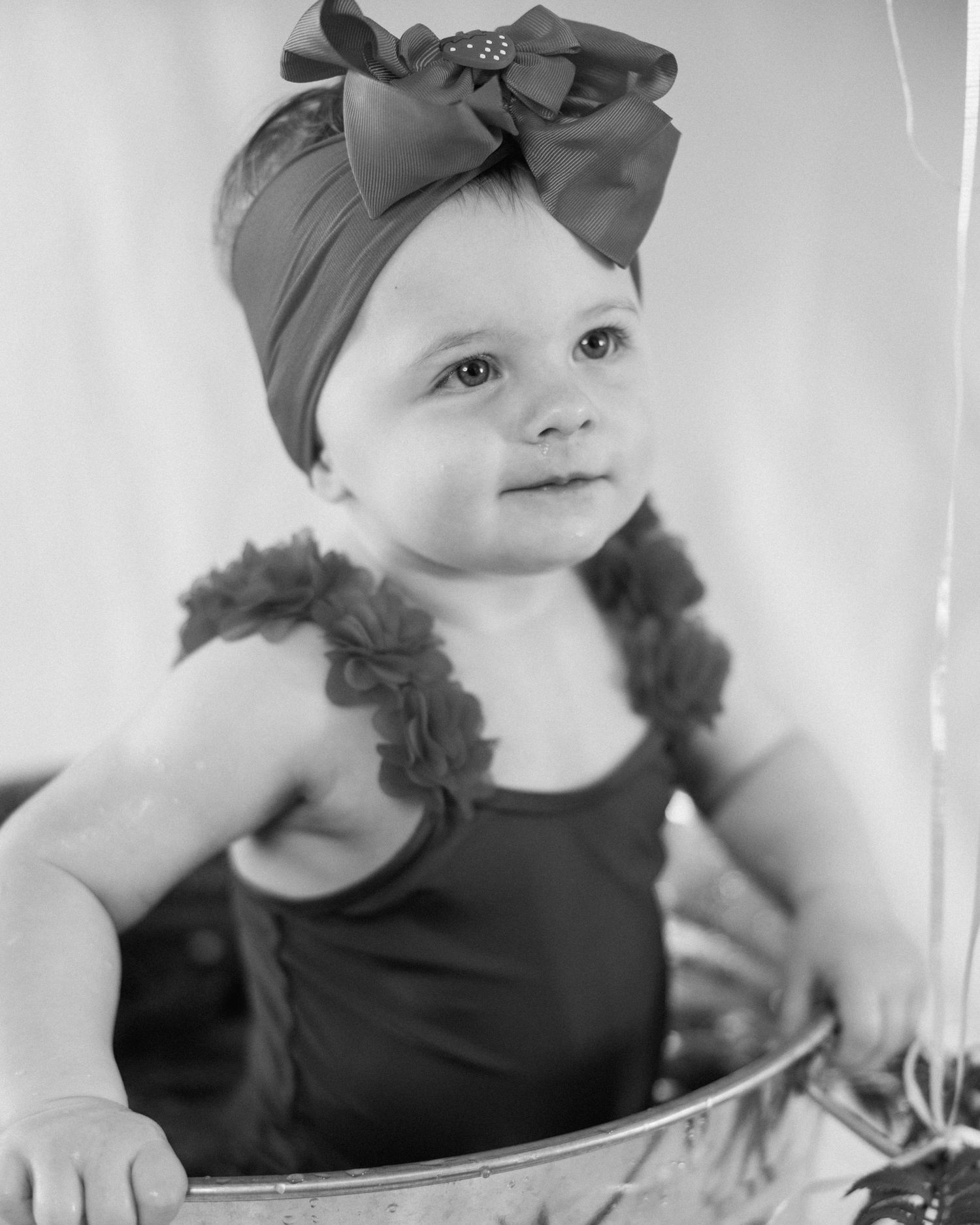 Black and white portrait of a cute baby girl wearing a bow headband and ruffled romper, sitting in a vintage tub.