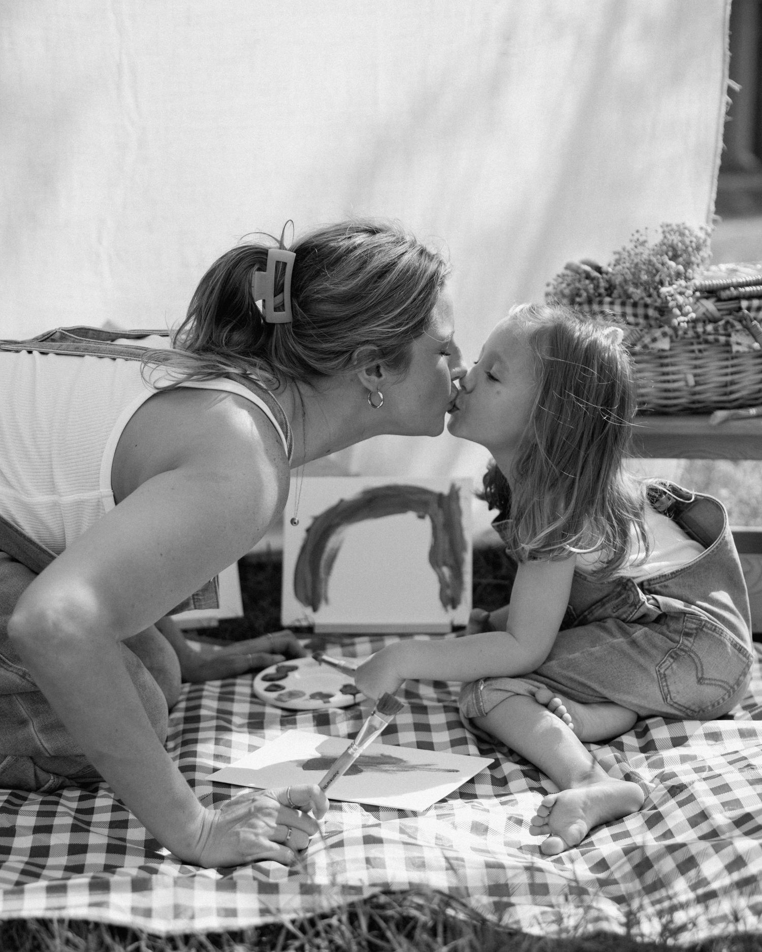 Black and white photo of a mother and young daughter sharing a kiss during an outdoor picnic with painting supplies.