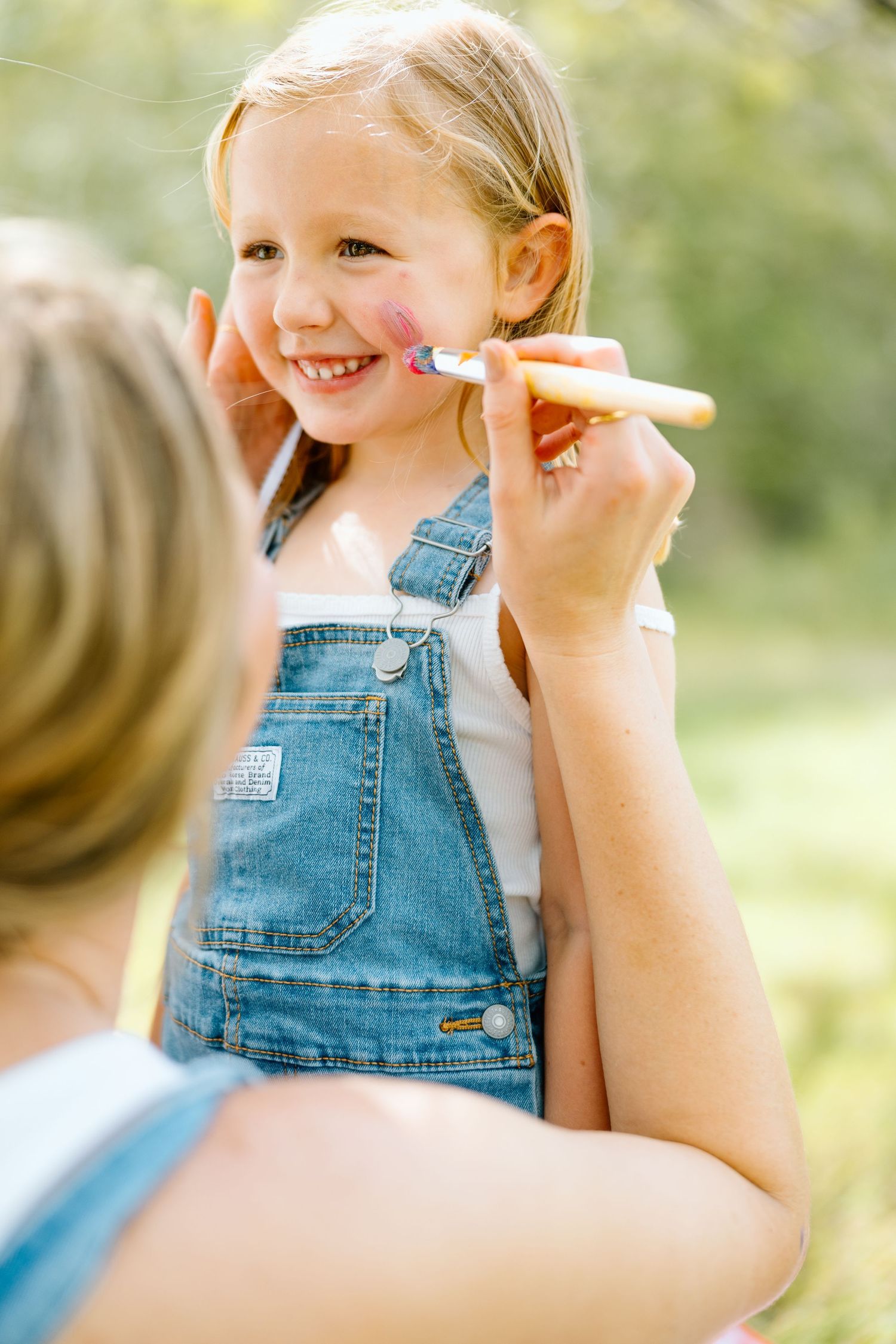 A smiling girl in denim overalls gets her face painted with pink design outdoors on a sunny day.