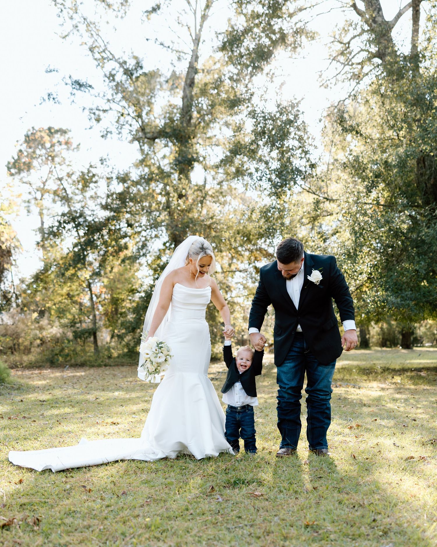 Bride and groom in wedding attire walk with toddler on sunny lawn surrounded by trees.
