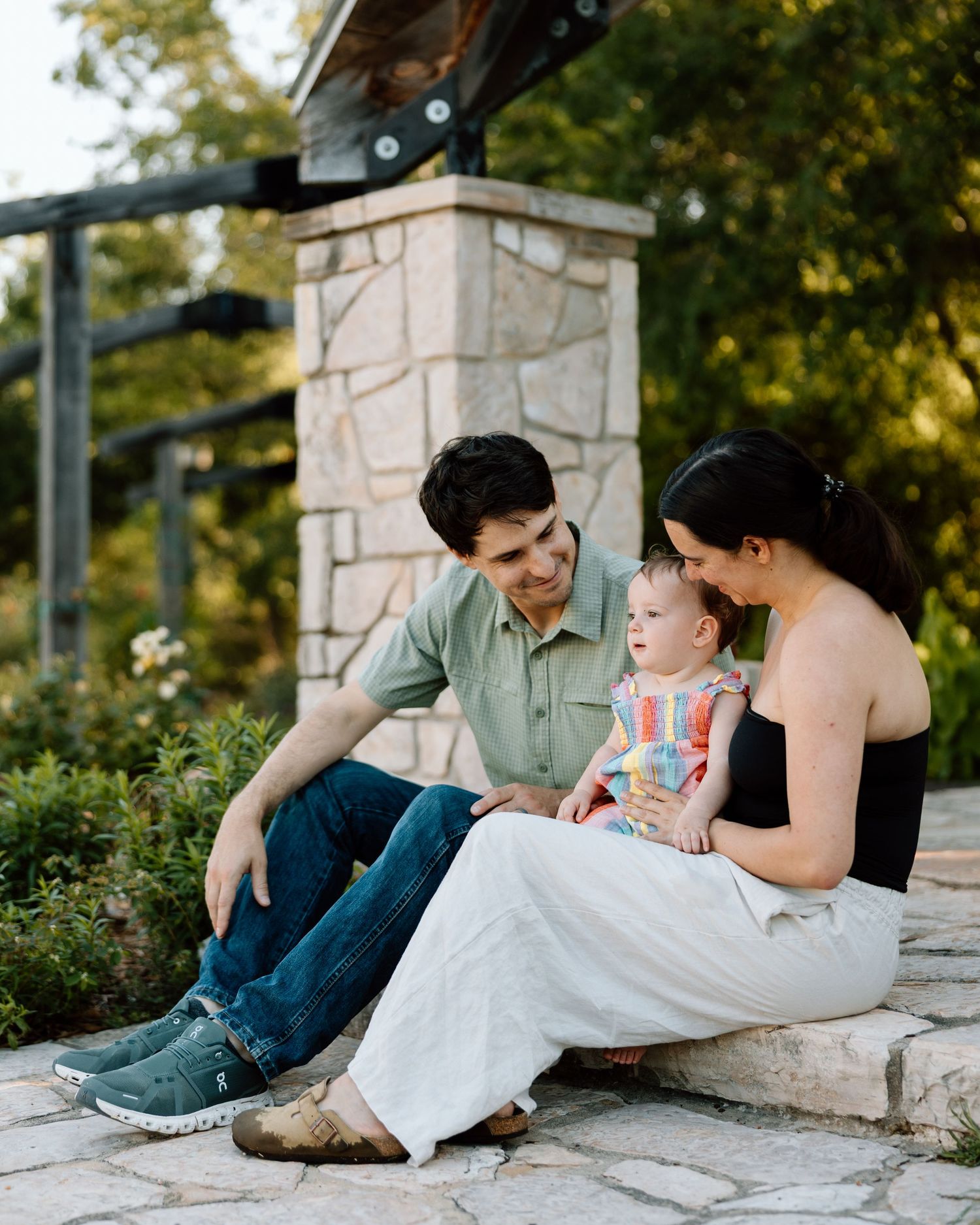 A young family sits together on stone steps outdoors, parents smiling at their baby in a colorful outfit.