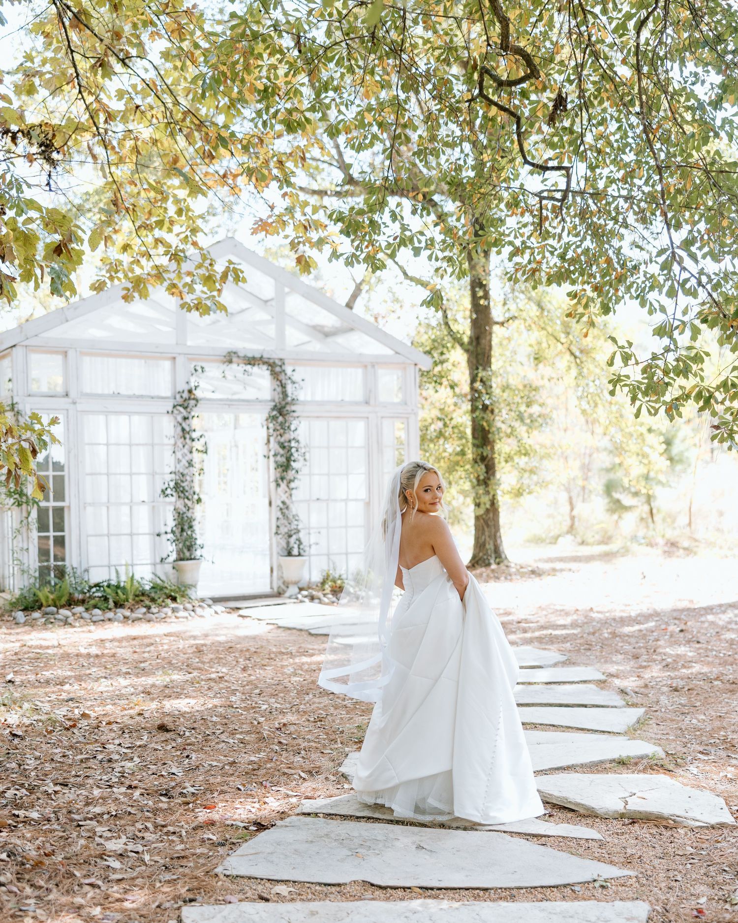 Bride in strapless white gown with flowing skirt walks stone path toward white greenhouse chapel surrounded by trees.
