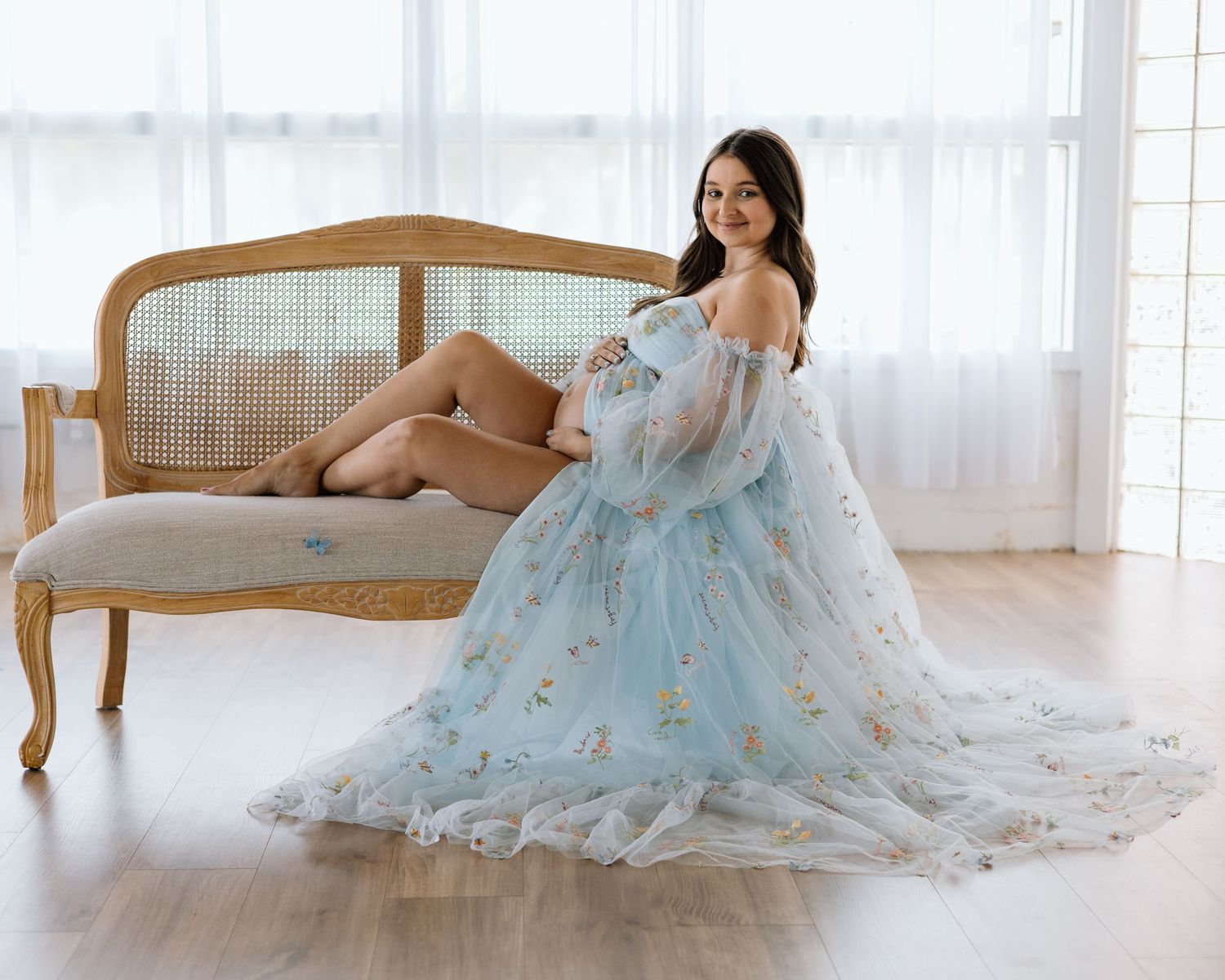 Pregnant woman in a flowy light blue floral tulle maternity gown posed elegantly on a vintage chaise lounge.