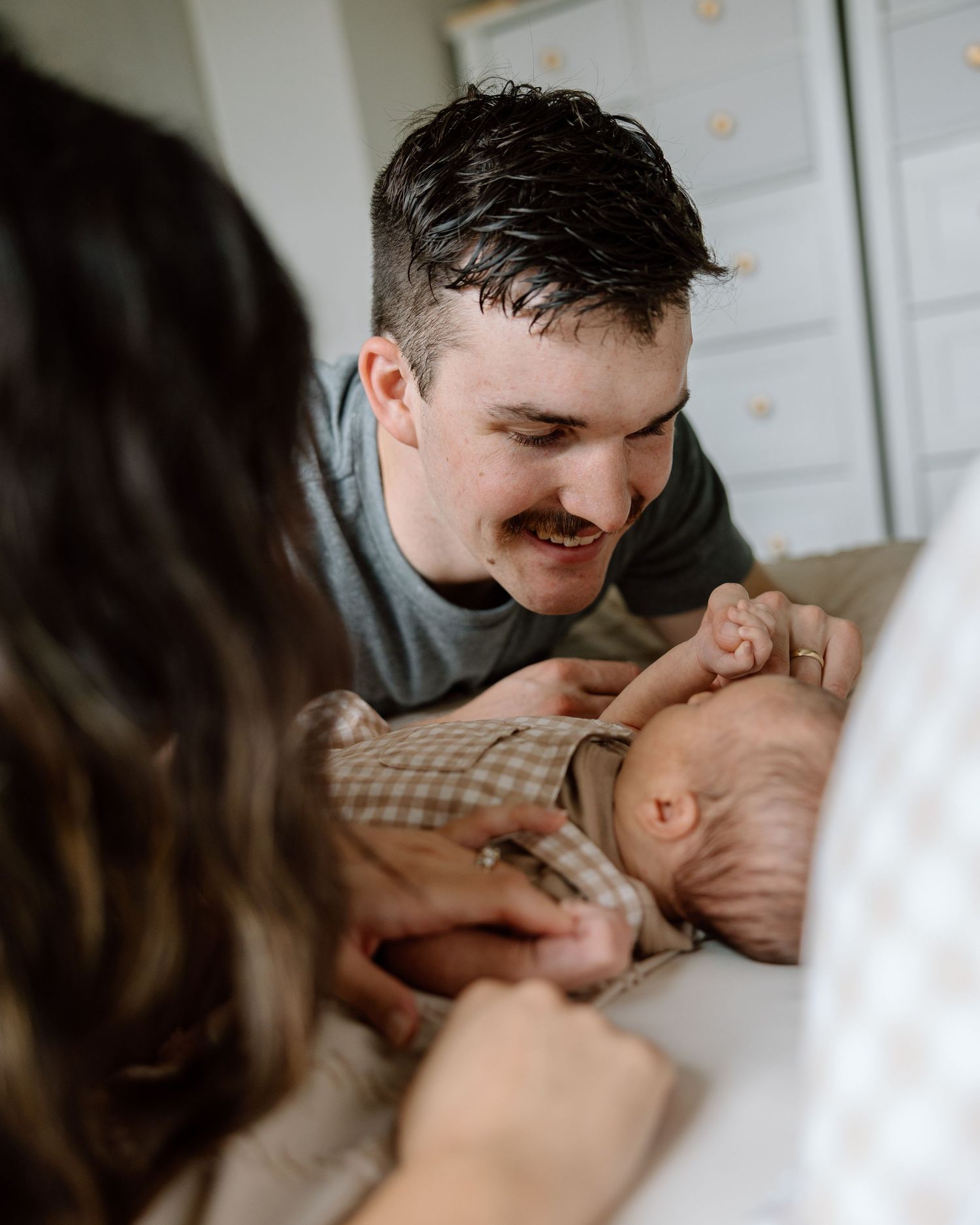 A smiling father with a mustache leans over a newborn baby while a mother looks on during an intimate family photo session.
