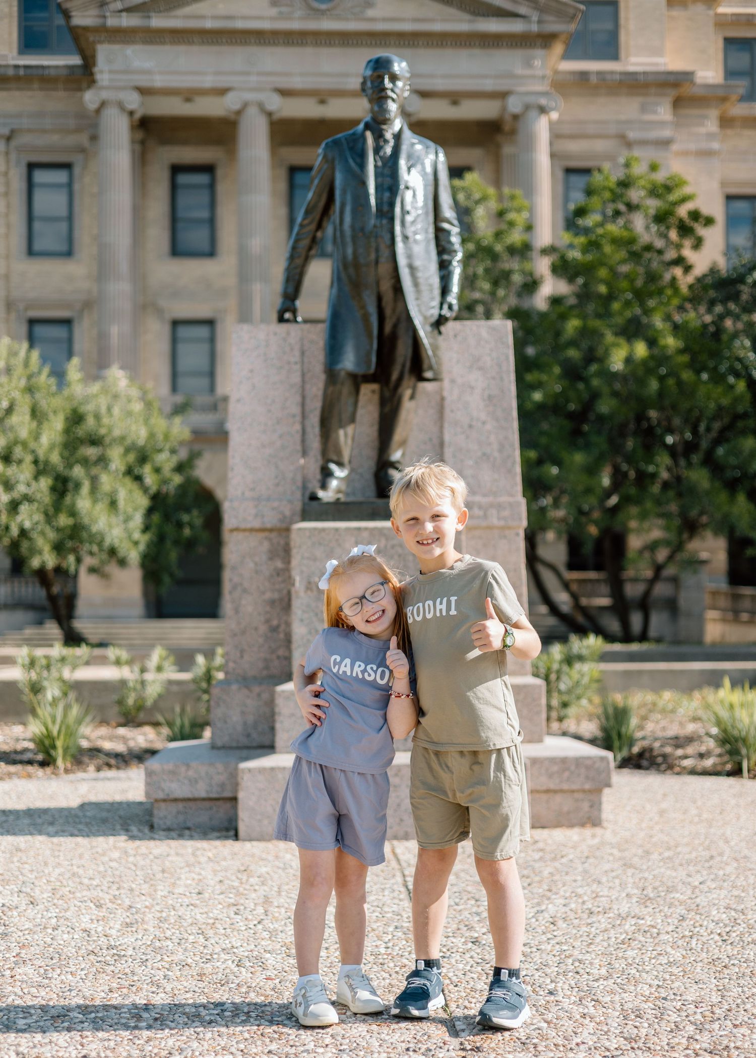 Two young children in matching gray shirts smile in front of a bronze statue outside a historic building.