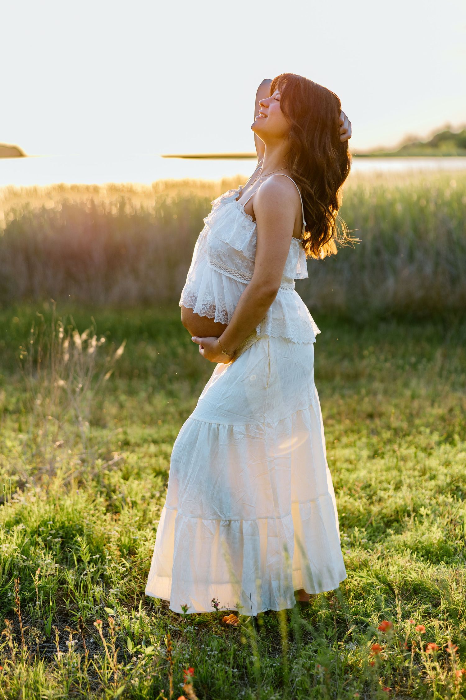 Pregnant woman in white lace dress holding belly in sunlit field at golden hour.