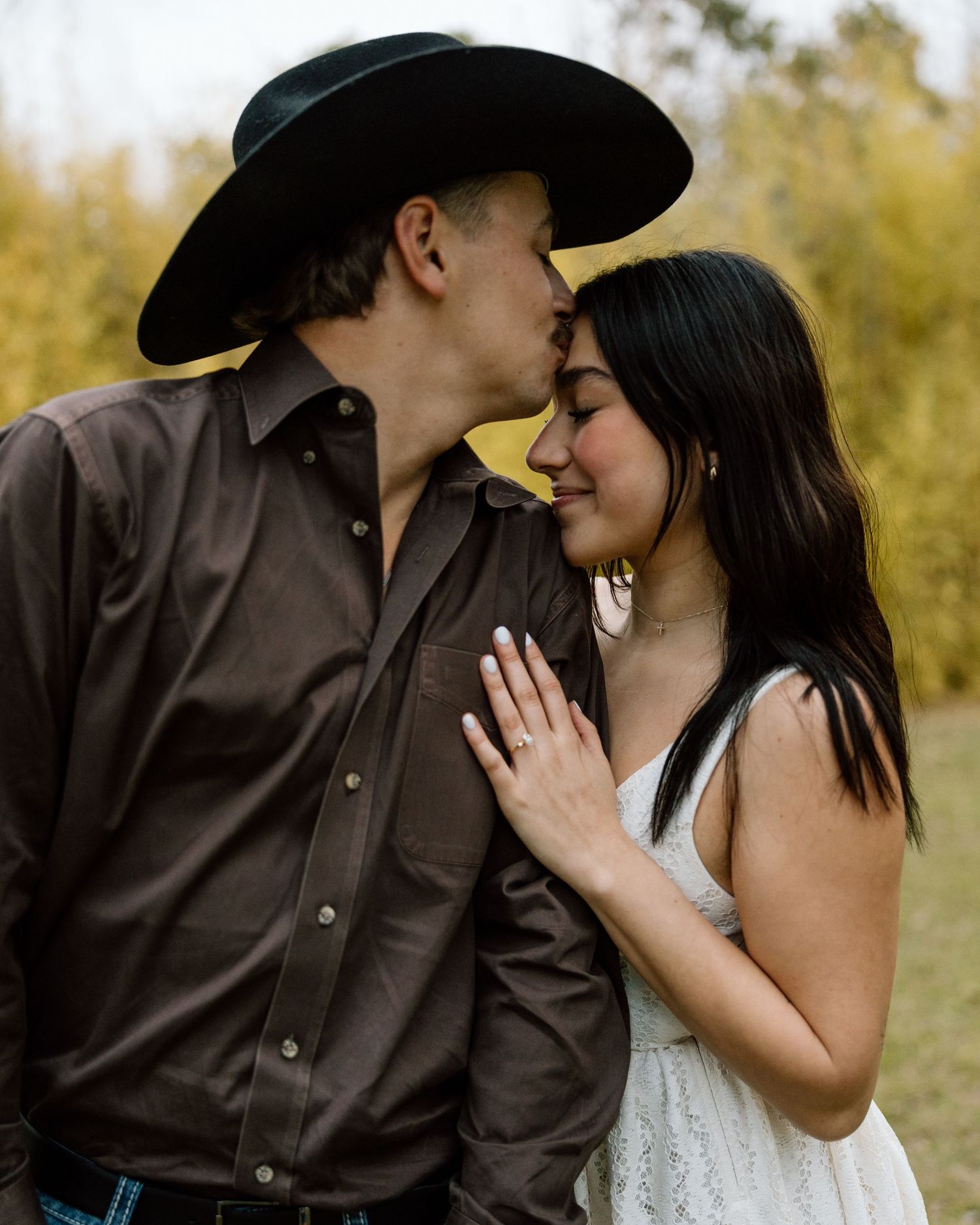Man in brown shirt kissing womans forehead while she displays diamond engagement ring outdoors.