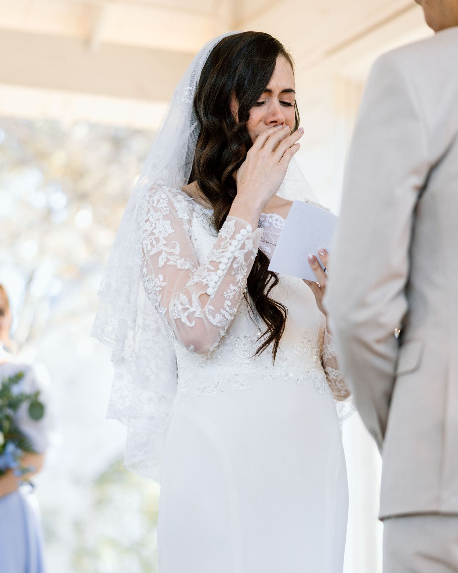 Emotional bride in lace wedding dress reading vows during outdoor ceremony.