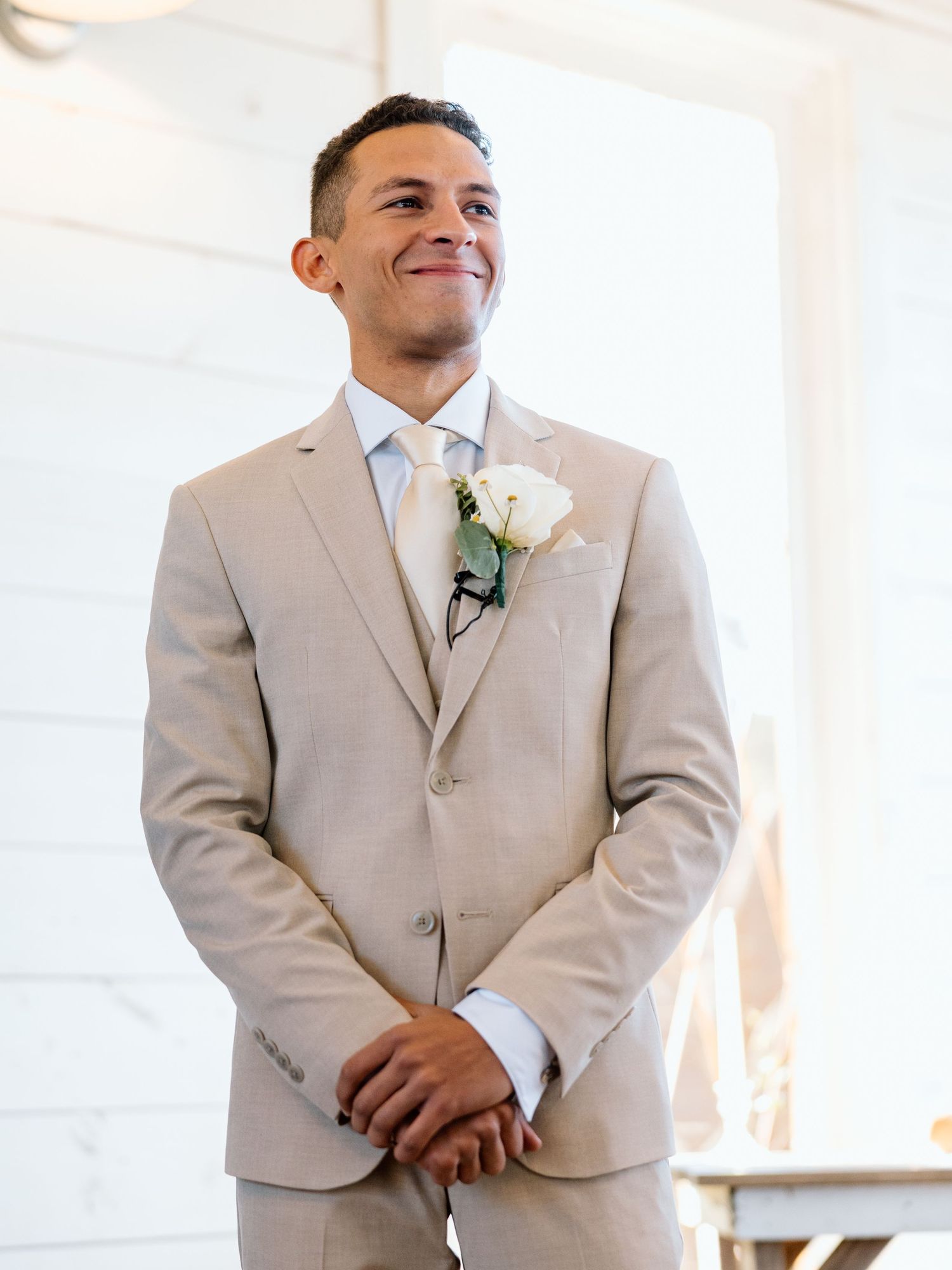 Groom in beige suit with white rose boutonniere smiling at wedding ceremony altar.