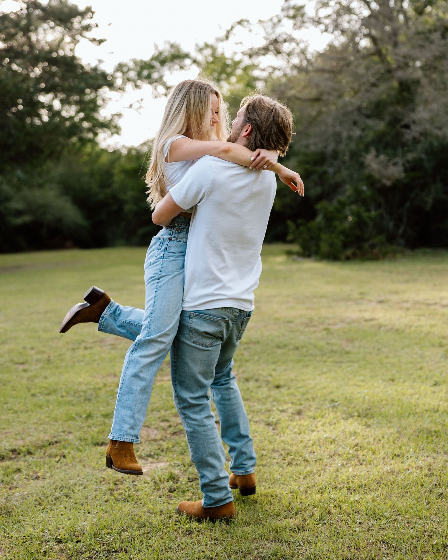 Couple embracing and kissing in a green park, wearing jeans and brown boots, sharing a romantic outdoor moment.