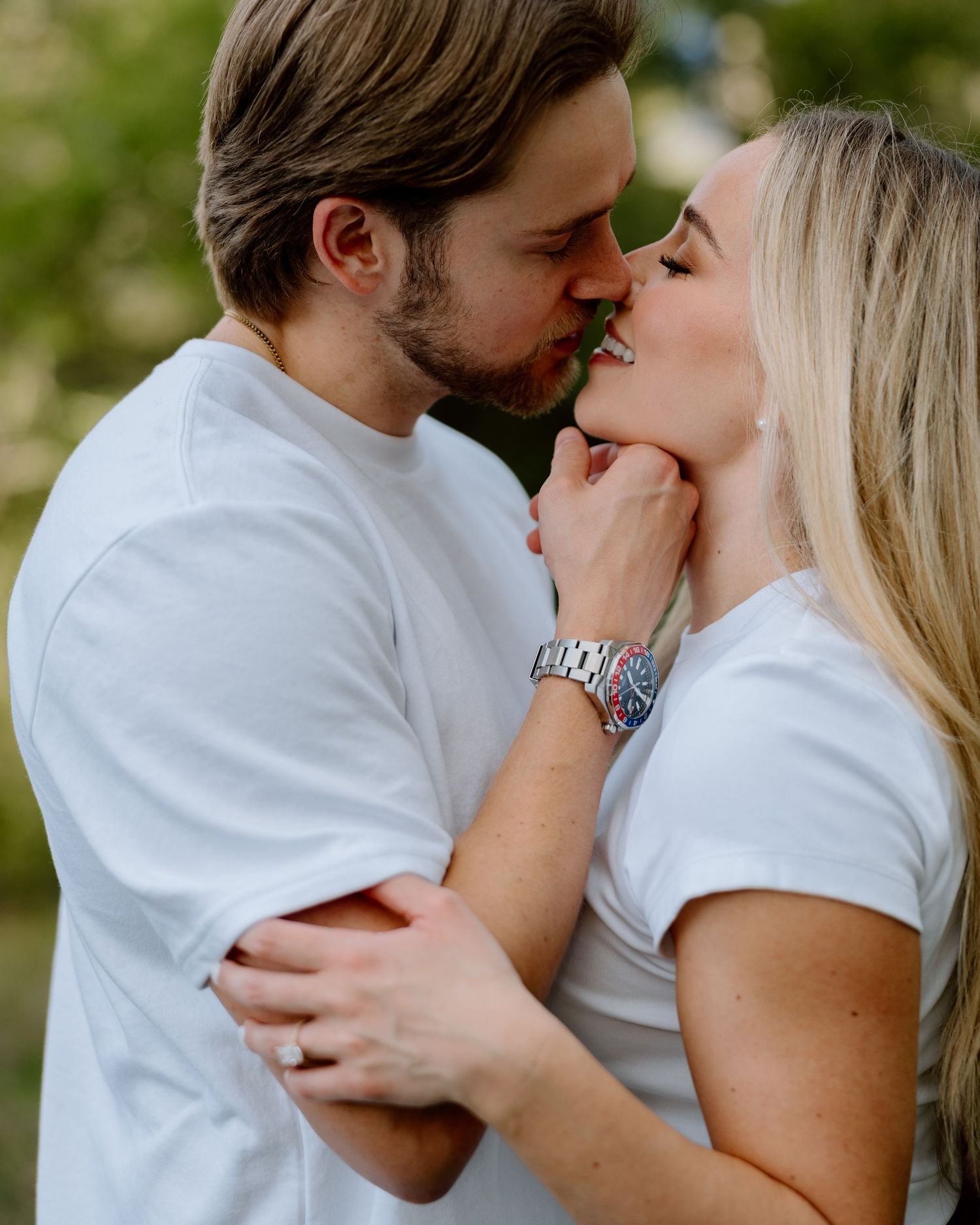 A couple in matching white t-shirts shares a romantic kiss outdoors, with the man gently holding the woman's face.
