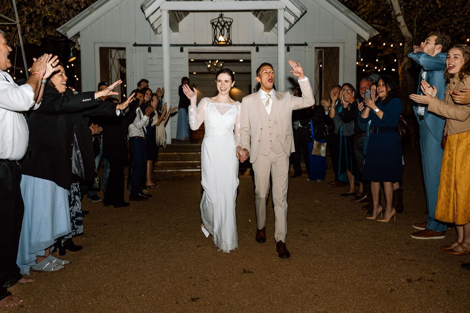 Newlyweds walk hand in hand through cheering guests during grand exit from white chapel at night.