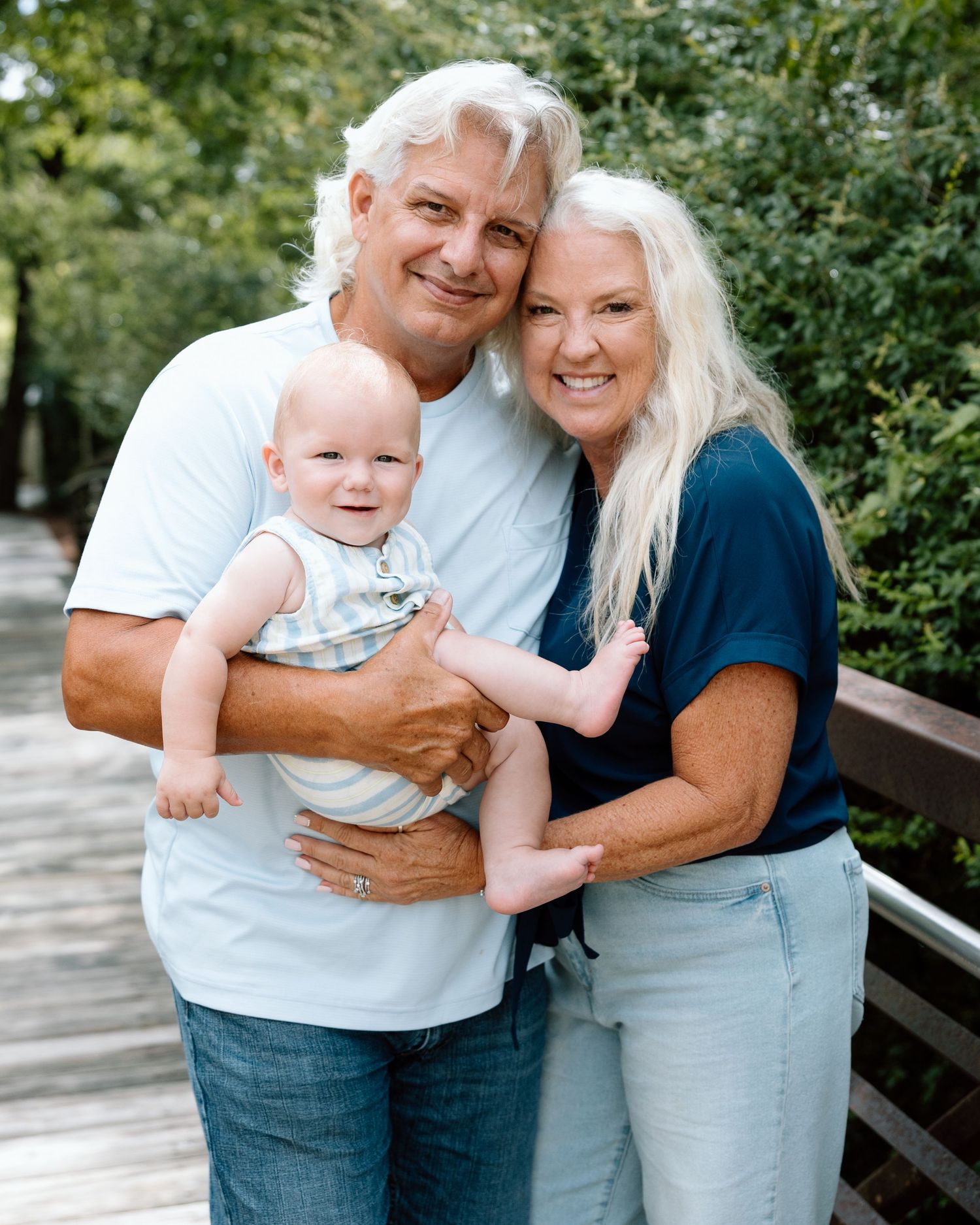 Smiling grandparents holding a happy baby on a wooden bridge surrounded by lush green trees.
