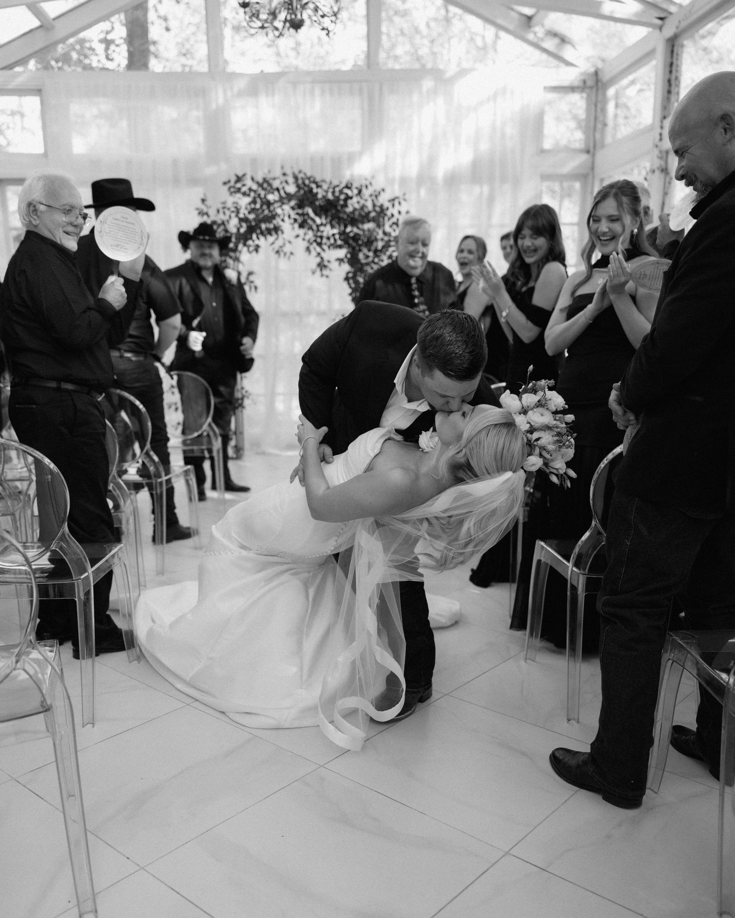 Black and white photo of groom dipping bride for a kiss at the altar as wedding guests cheer.