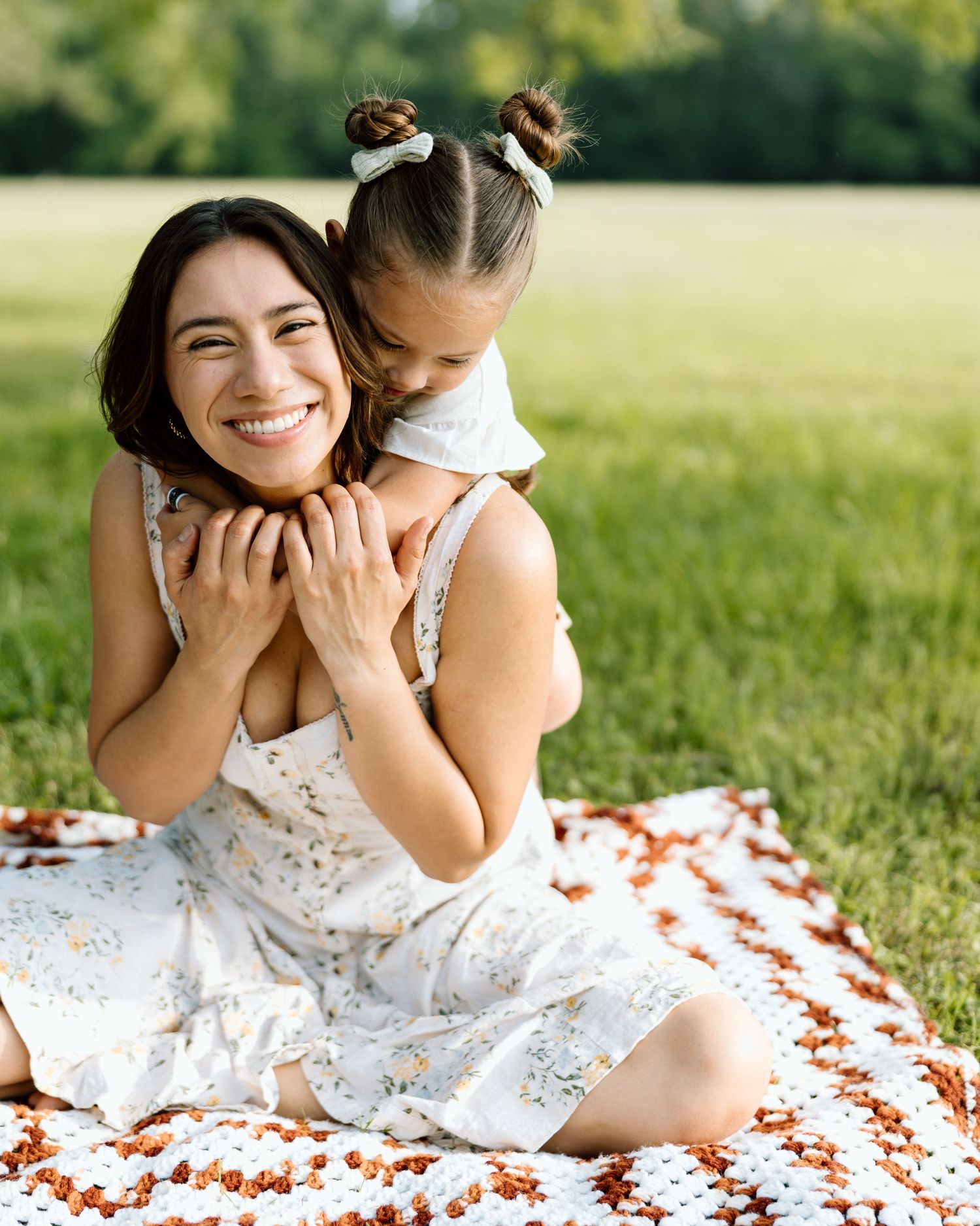 A smiling woman and young girl share a joyful hug on a blanket in a sunny green park.