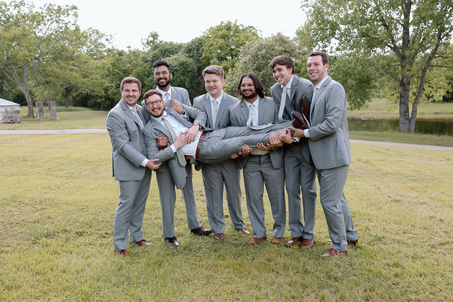 Groomsmen in matching grey suits playfully carry someone in a fun group photo.
